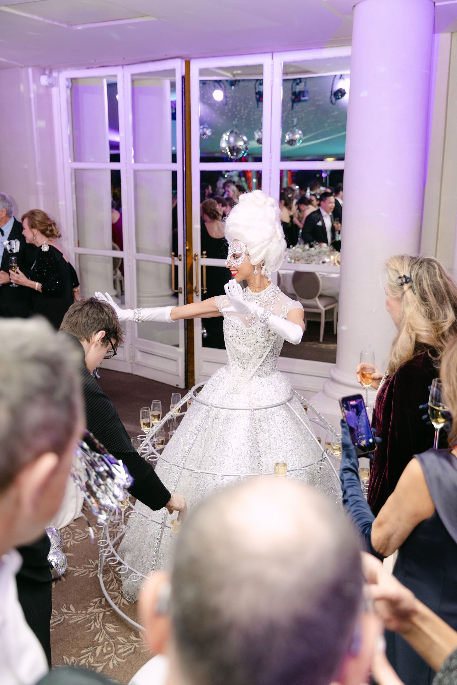 Glamorous entertainer in silver ball gown with hoop skirt serving champagne to guests at a formal event.