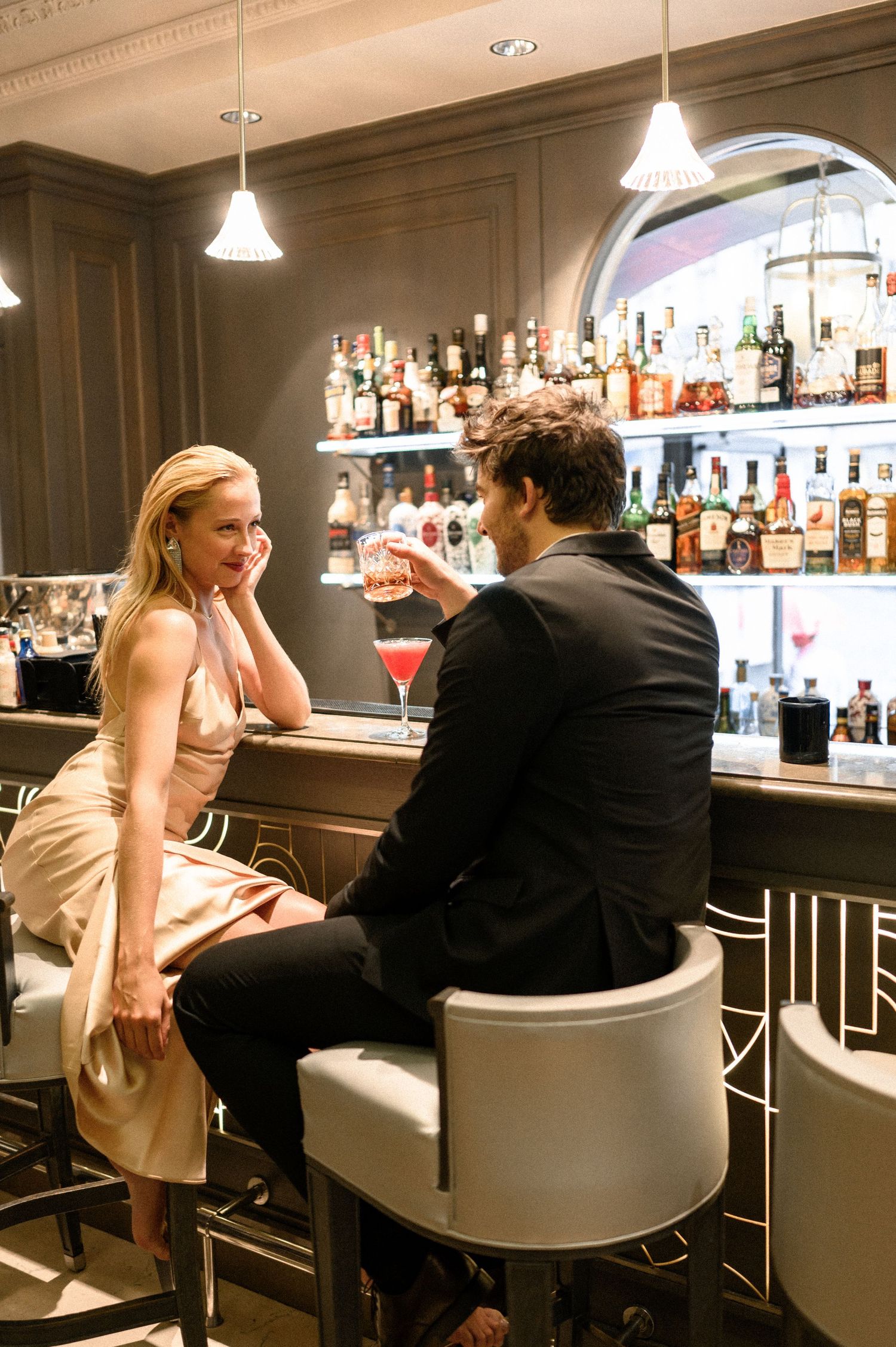 Stylish couple sharing cocktails at a sophisticated hotel bar with backlit liquor shelves.