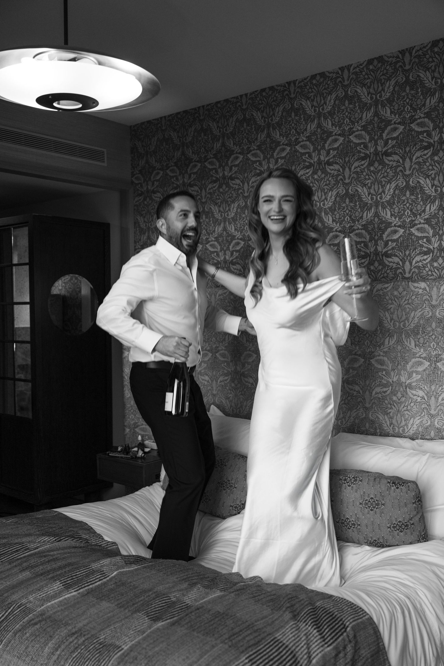 Black and white photo of joyful couple jumping on hotel bed, bride in white gown holding champagne.