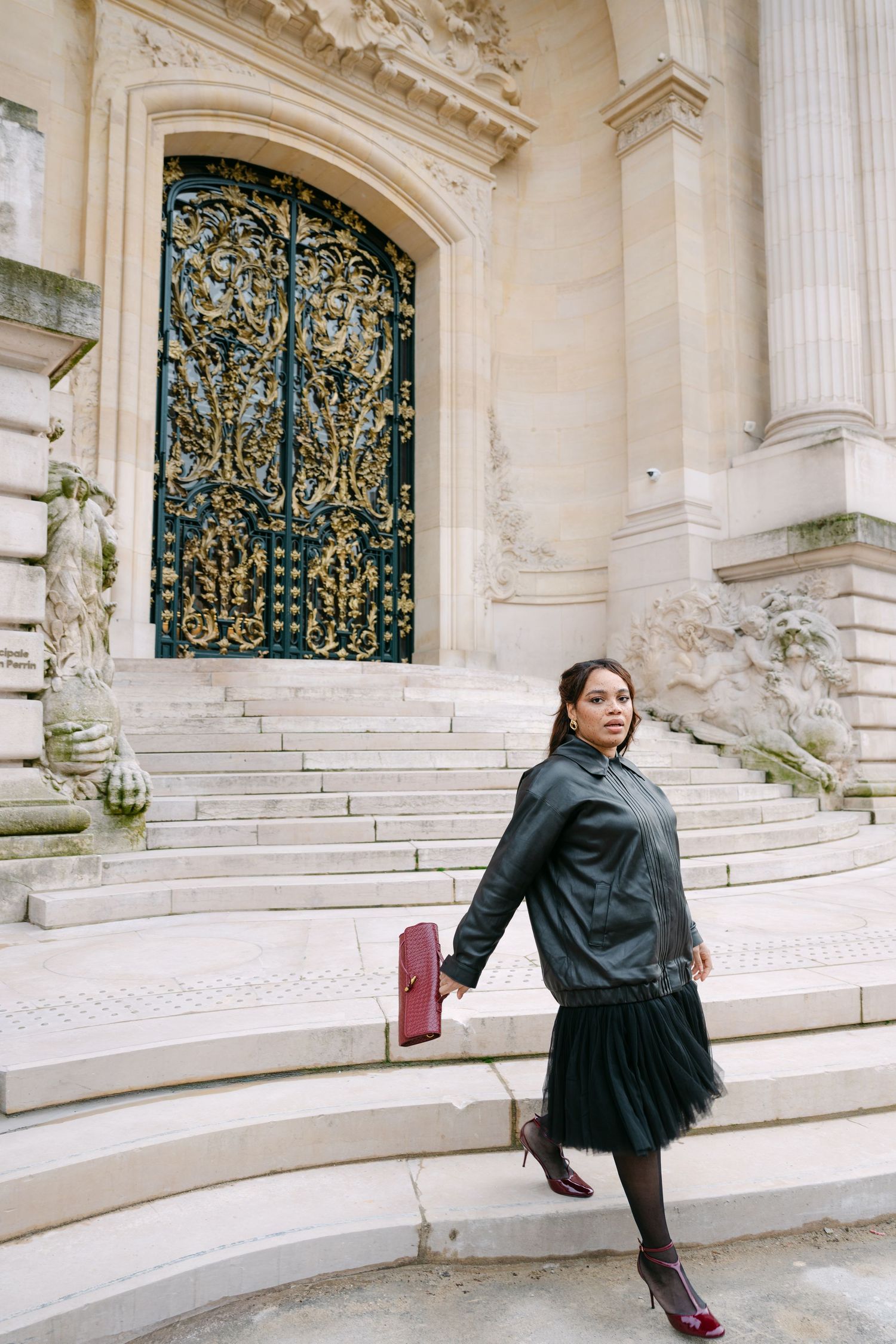 Woman in black leather jacket and tulle skirt poses on steps of ornate Parisian building with golden doors.