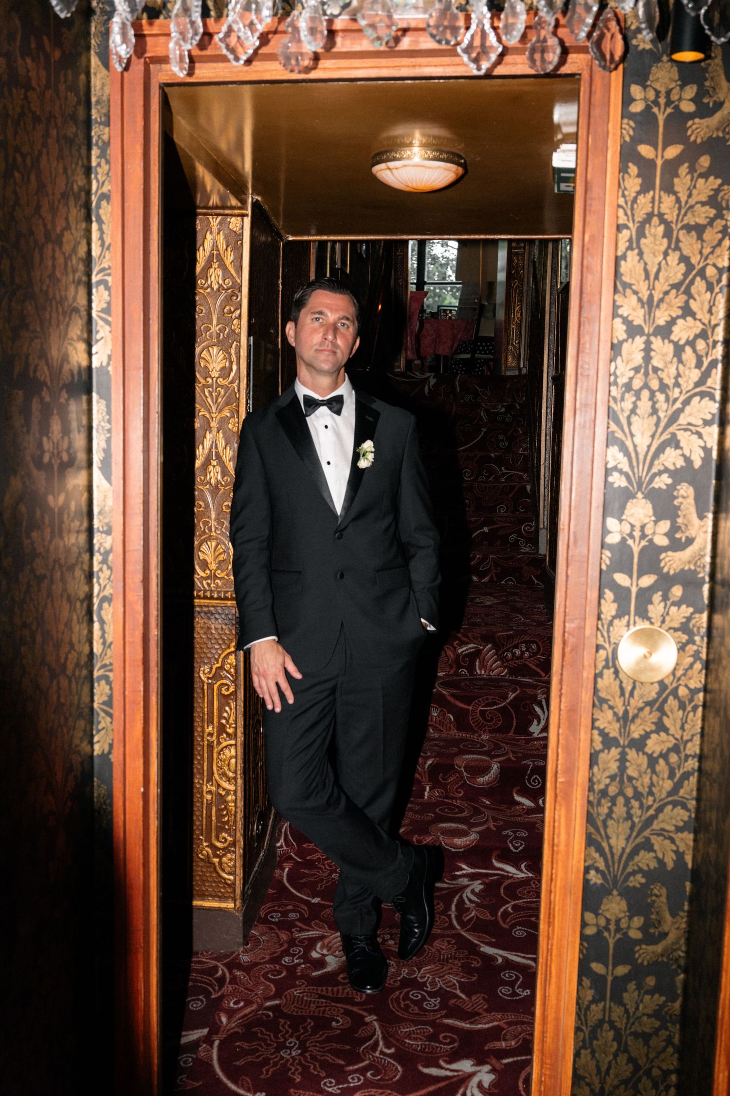 Groom in black tuxedo standing confidently in an ornate doorway with gold wallpaper.
