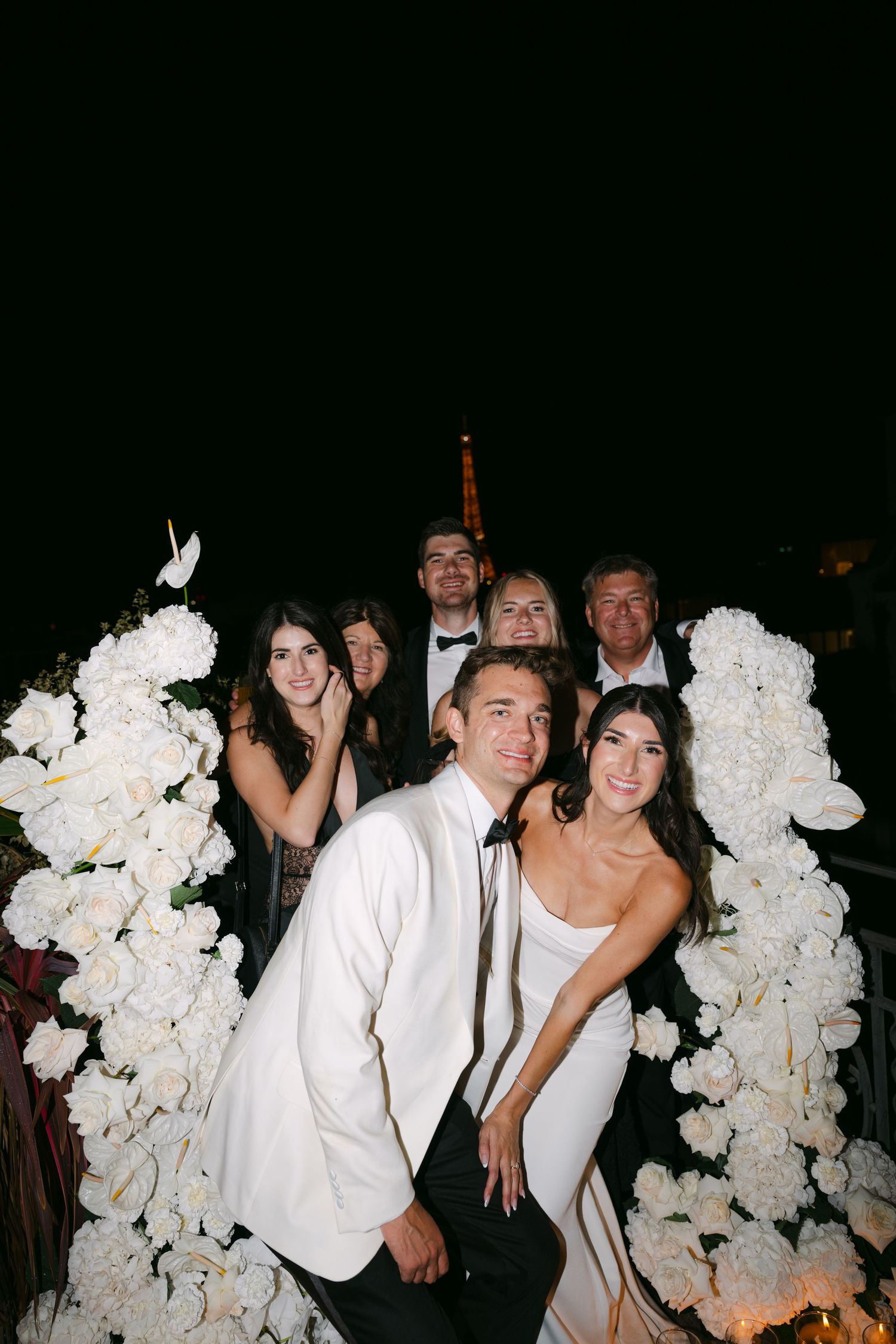 Wedding guests in formal attire gather around elegant white floral arrangements at an evening celebration.