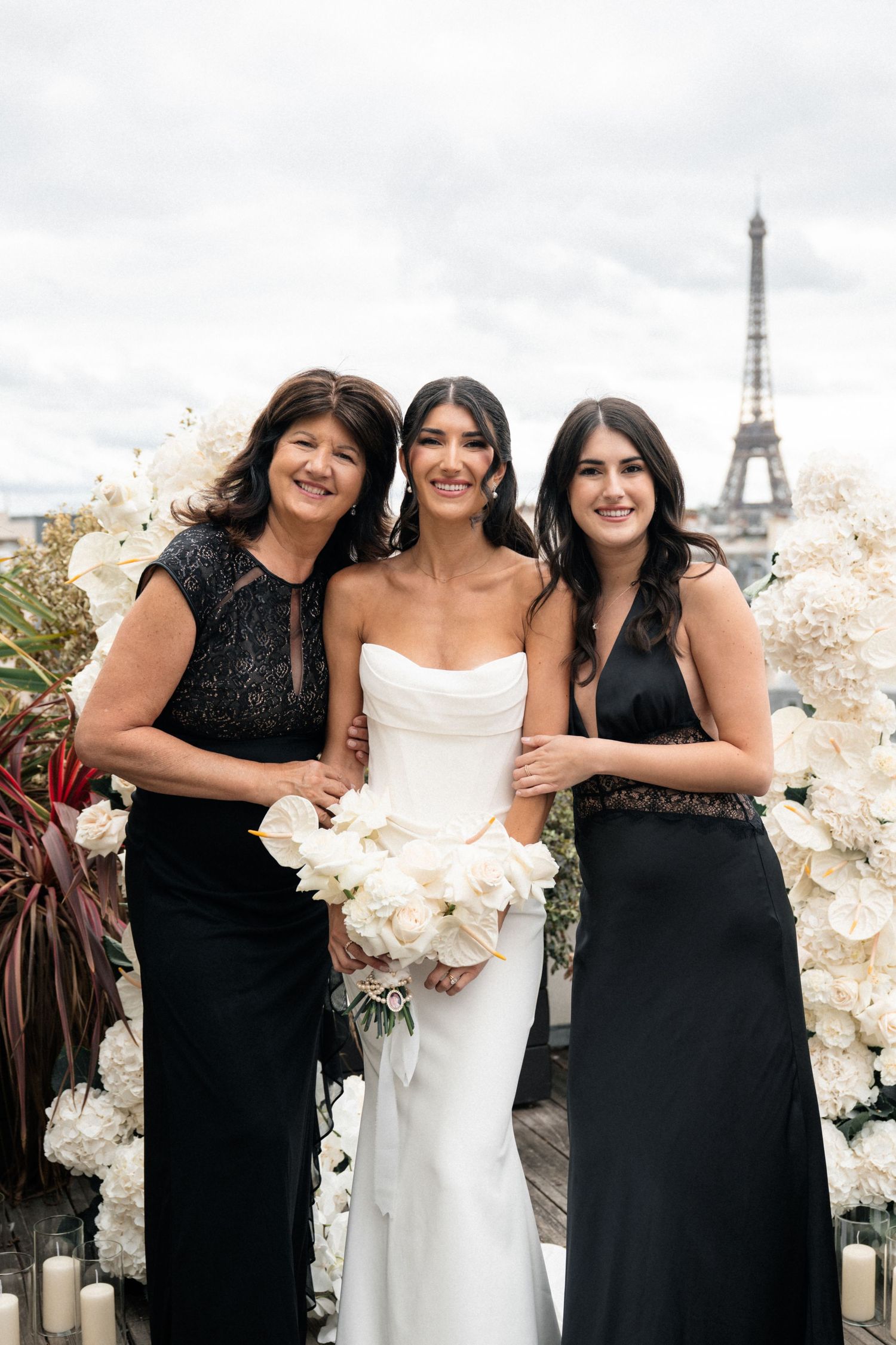 Three women in formal attire pose together at a rooftop venue in Paris with the Eiffel Tower visible in the background.