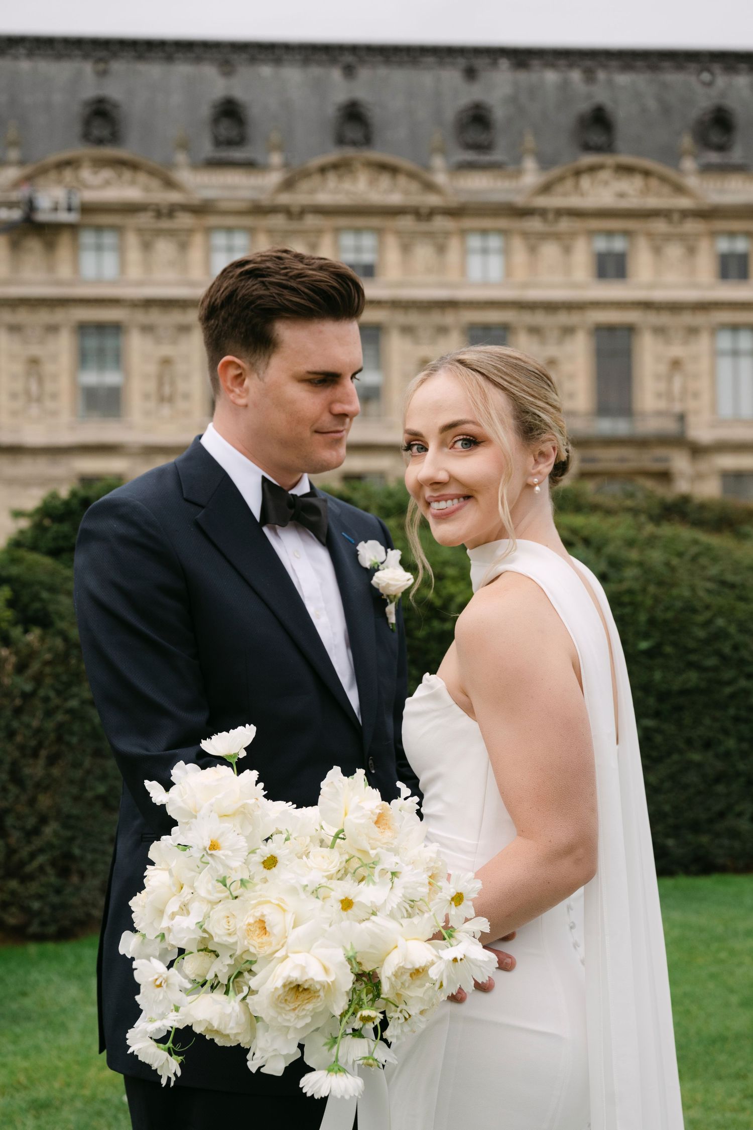 Elegant wedding couple poses with white floral bouquet in front of historic European mansion on manicured lawn.
