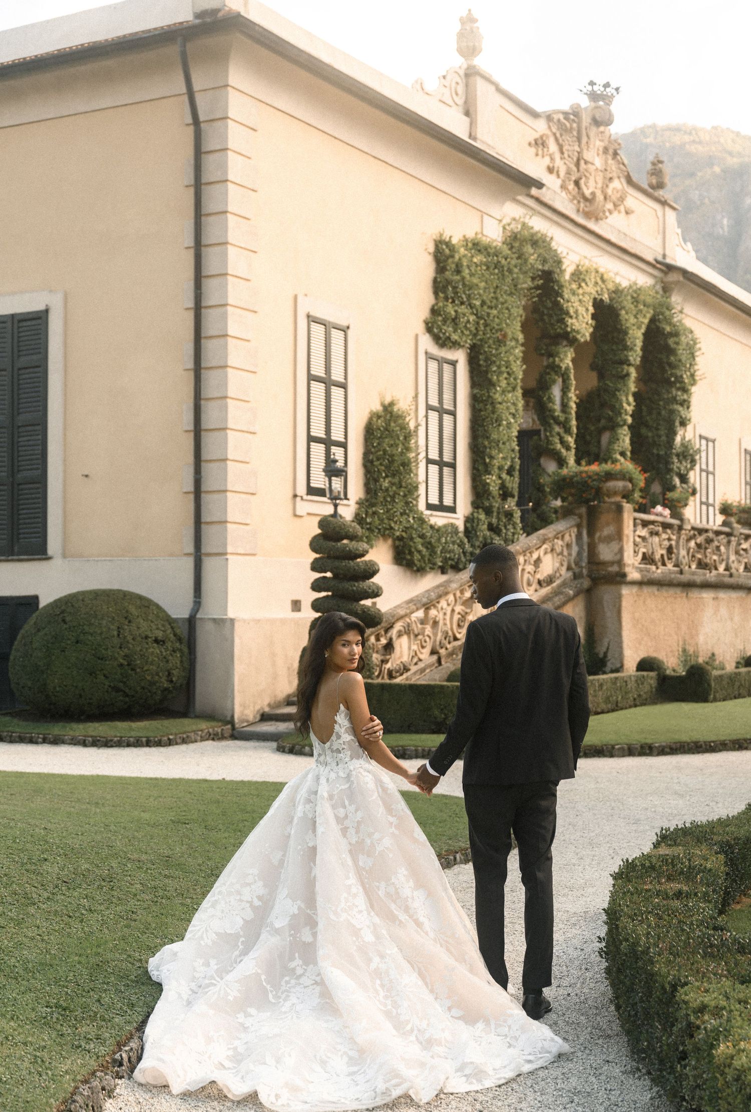 Bride and groom walking hand in hand outside a classic Italian villa with manicured gardens.