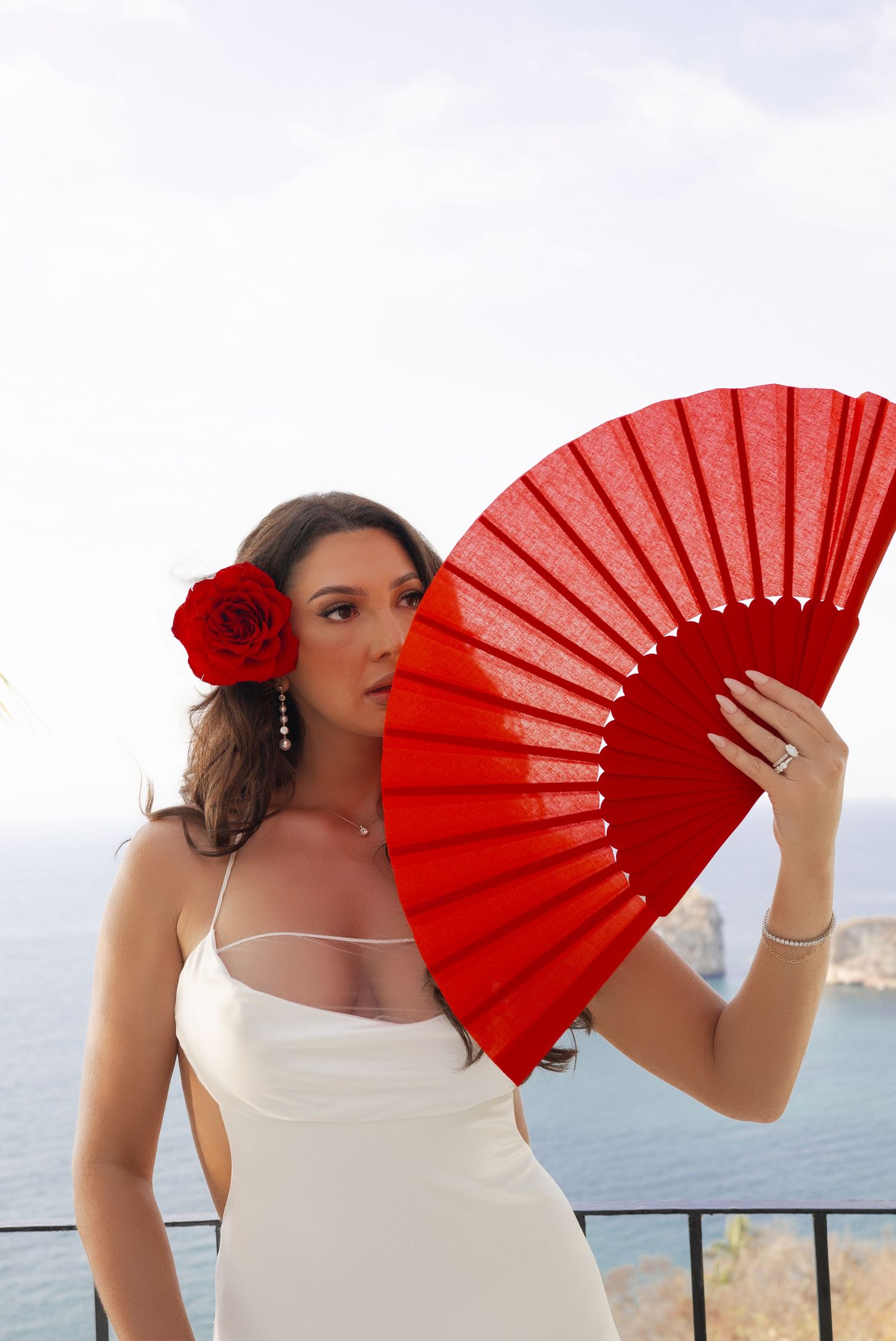 Woman in white dress holding red fan with rose in hair, posing against ocean backdrop.