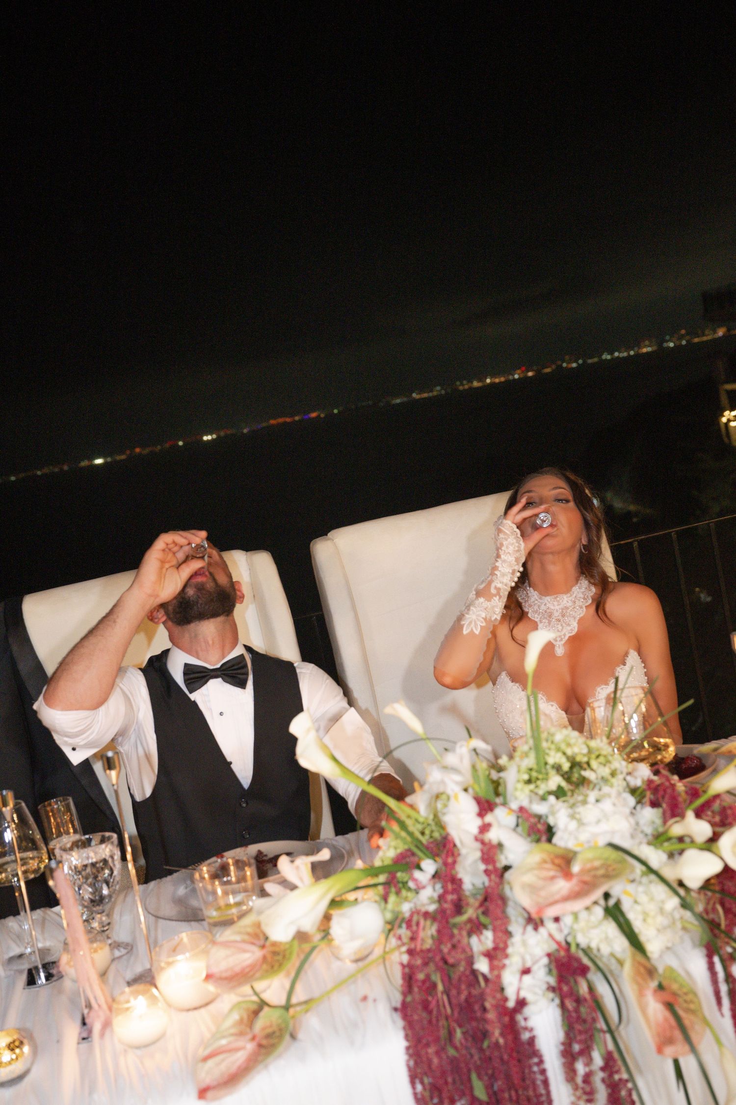Bride and groom laugh joyfully at their candlelit wedding reception table with city lights glowing in the night background.
