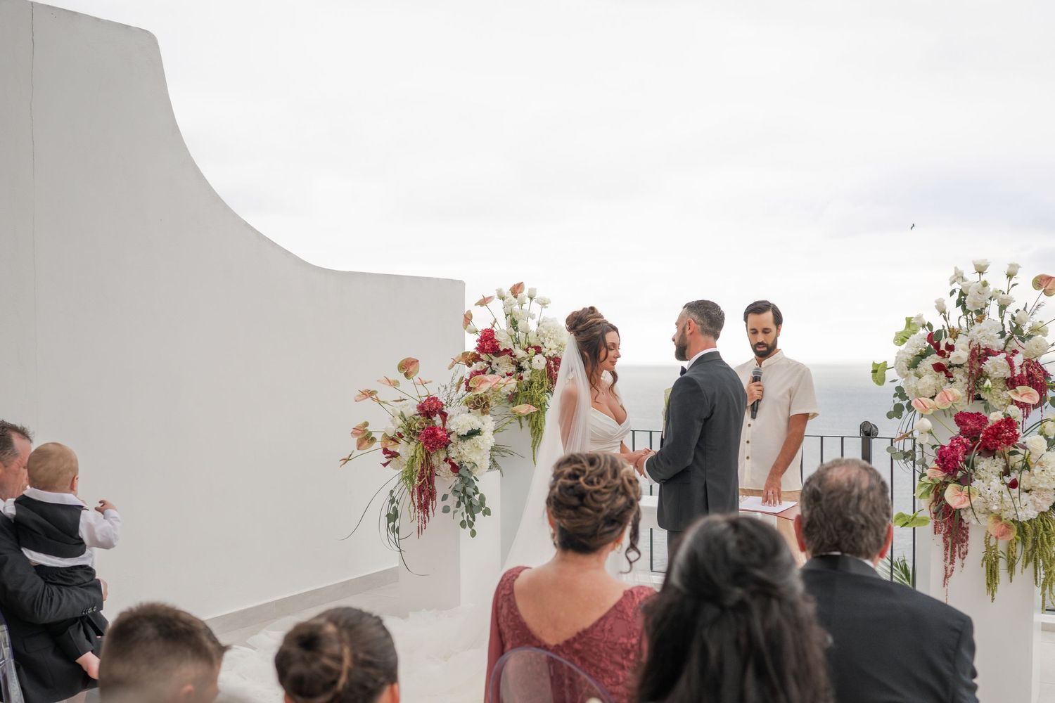 Bride and groom exchange vows at outdoor rooftop wedding ceremony with elegant red and white floral arrangements.