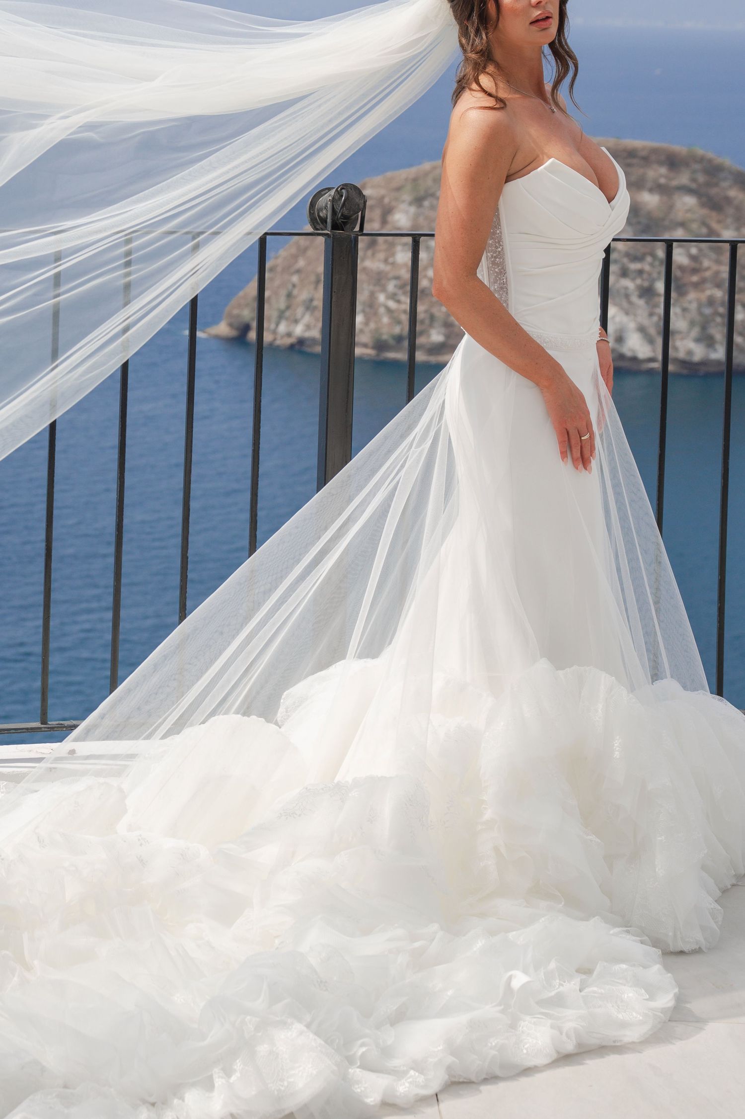 Bride in strapless white mermaid gown with flowing veil stands on balcony overlooking blue sea.