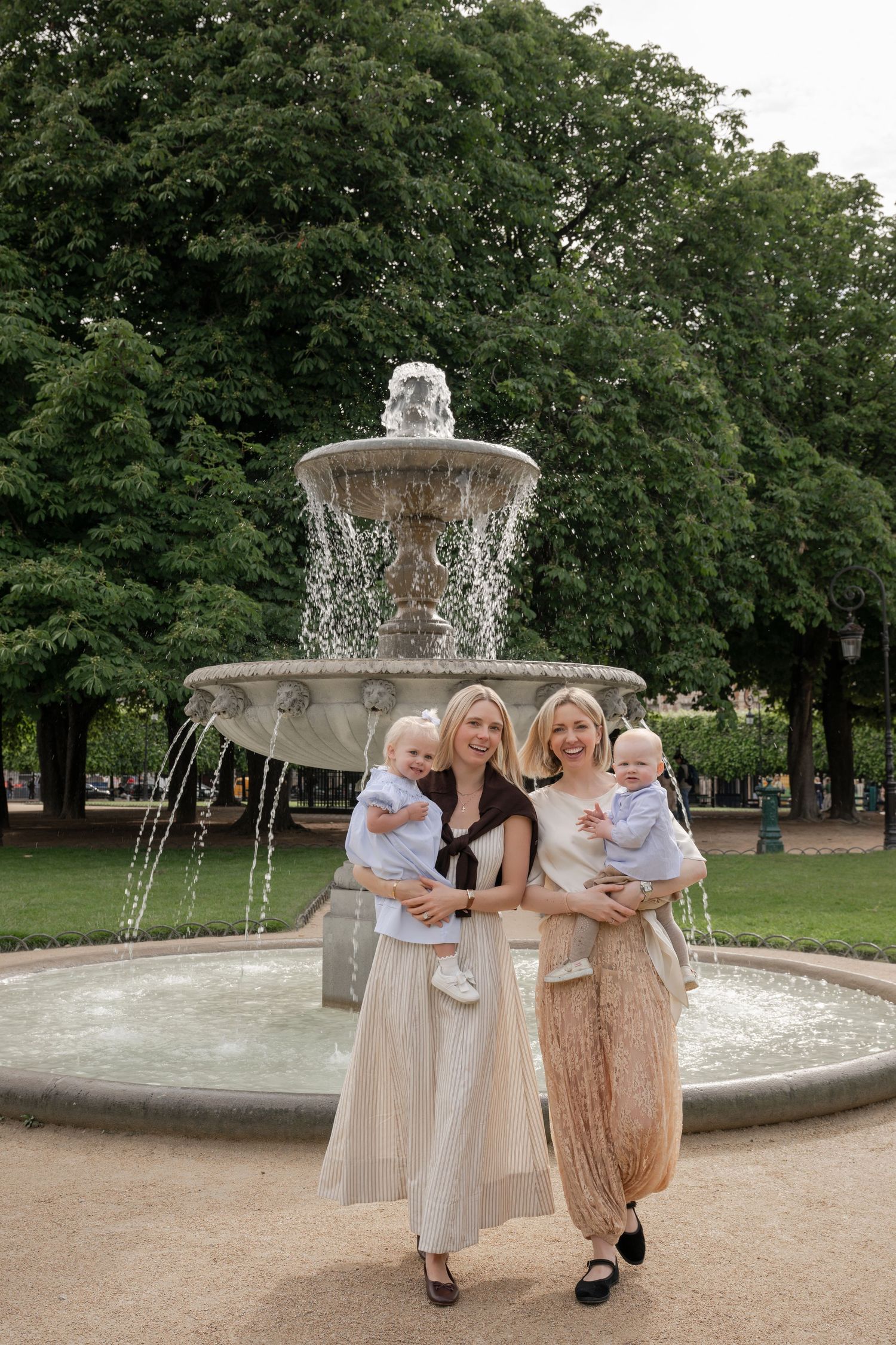 Two blonde women holding toddlers pose in front of an ornate stone fountain in a lush green park.