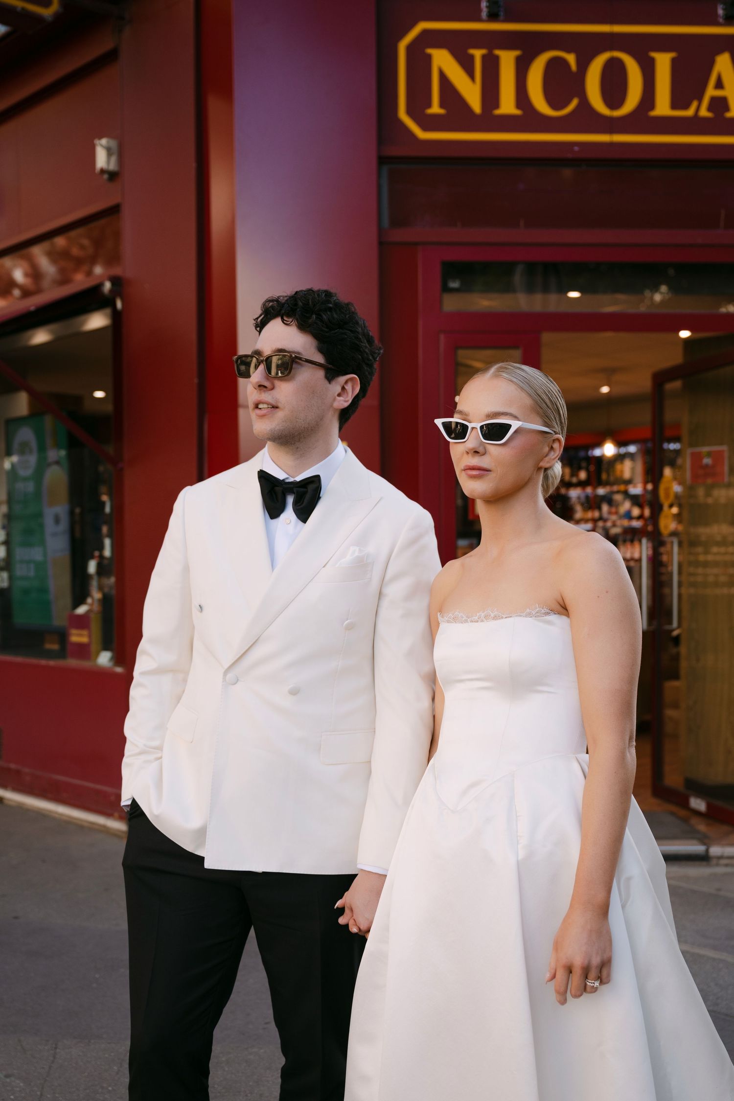 Stylish wedding couple in white outfits and sunglasses posing outside Nicolas wine shop in Paris.
