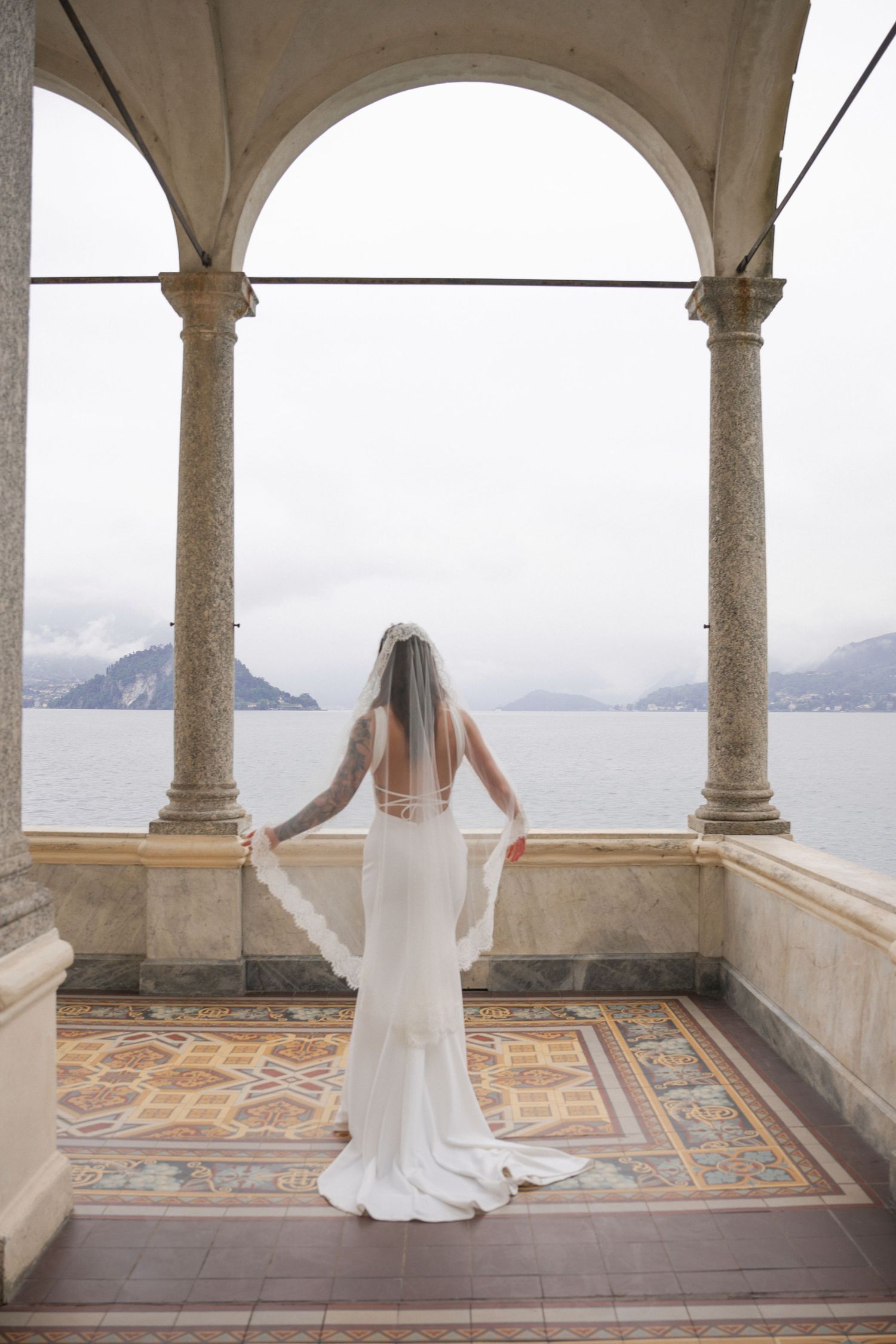 Bride in white gown and veil stands between marble columns of Italian villa overlooking misty lake.
