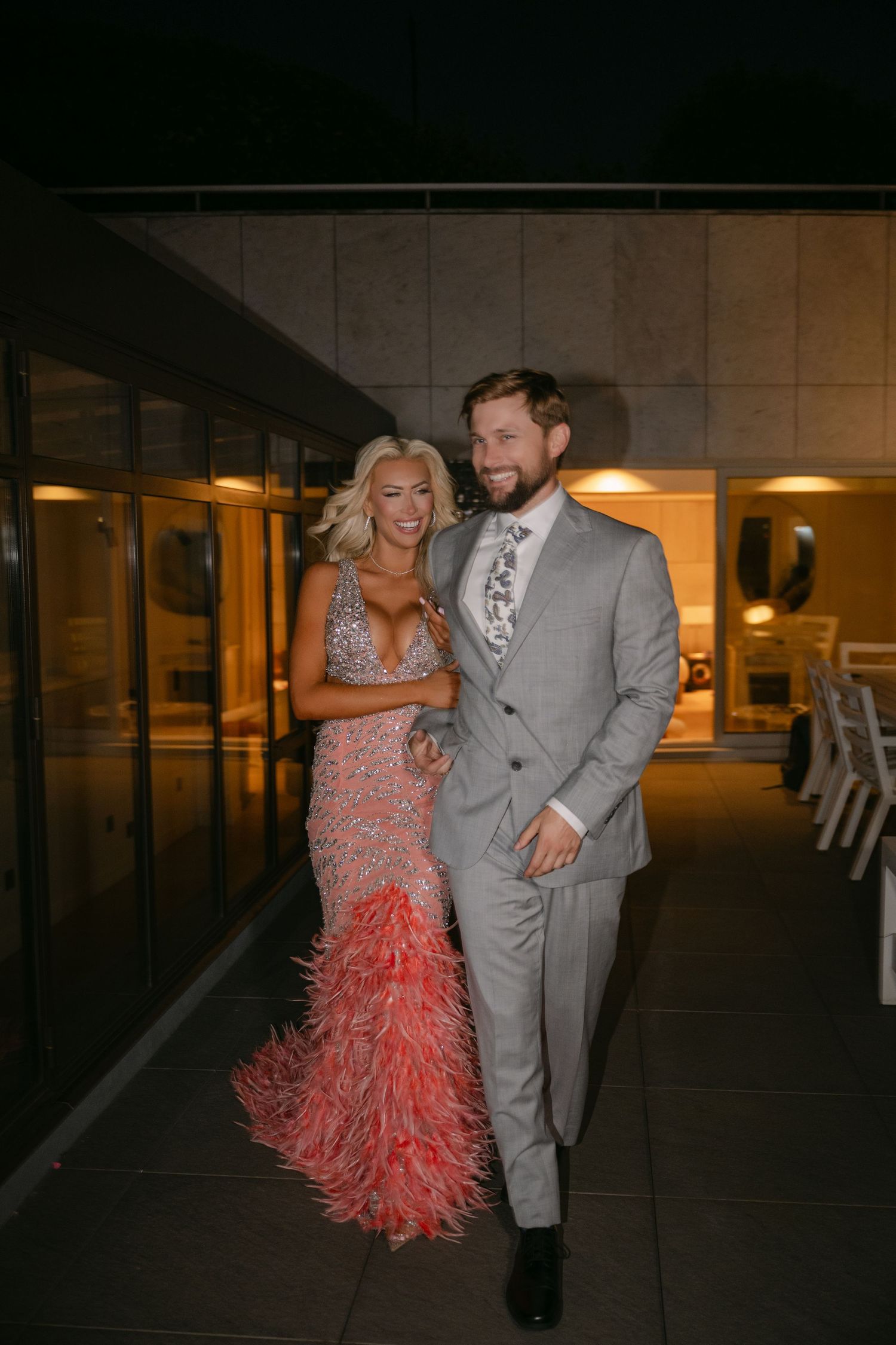 Couple posing outdoors at night, woman in pink feather gown and man in gray suit with floral tie.