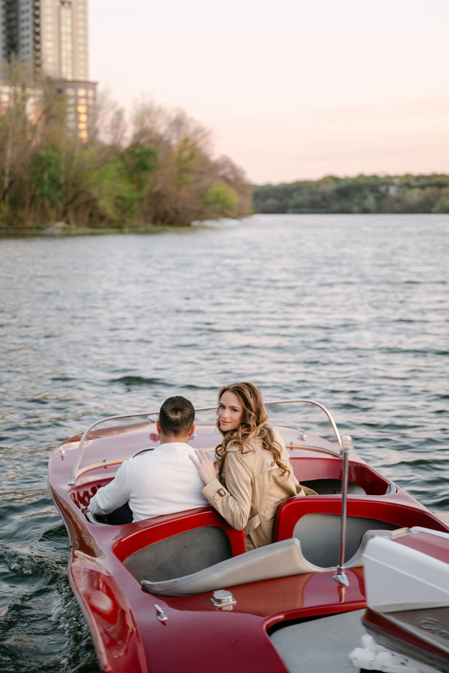 Couple sitting in a vintage red speedboat on a calm river at dusk.
