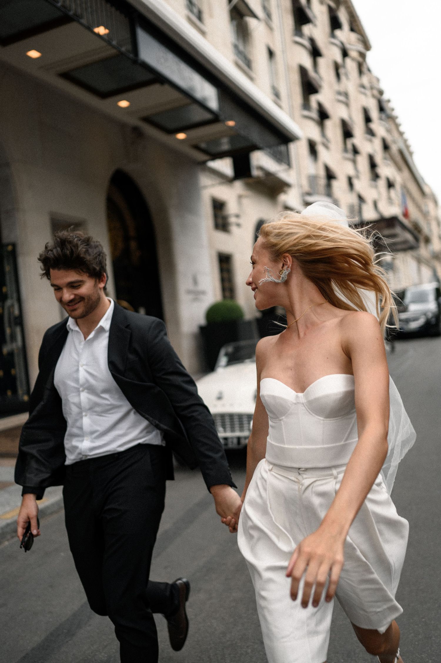 Joyful newlyweds running hand in hand on a Parisian street, bride in white strapless gown, groom in black suit.