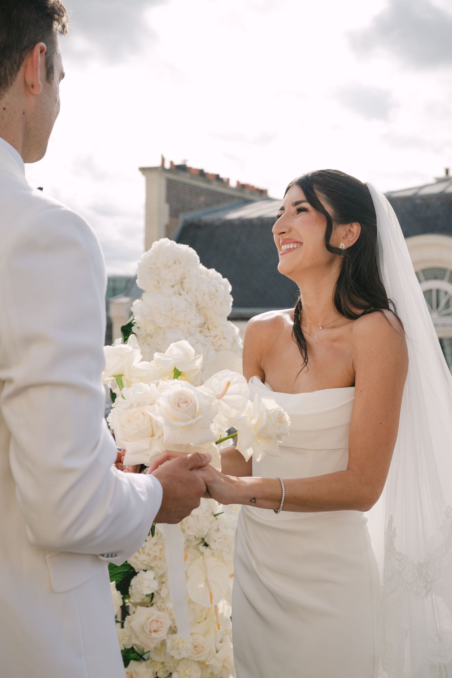 Radiant bride in white strapless dress and veil holds hands with groom surrounded by white roses.