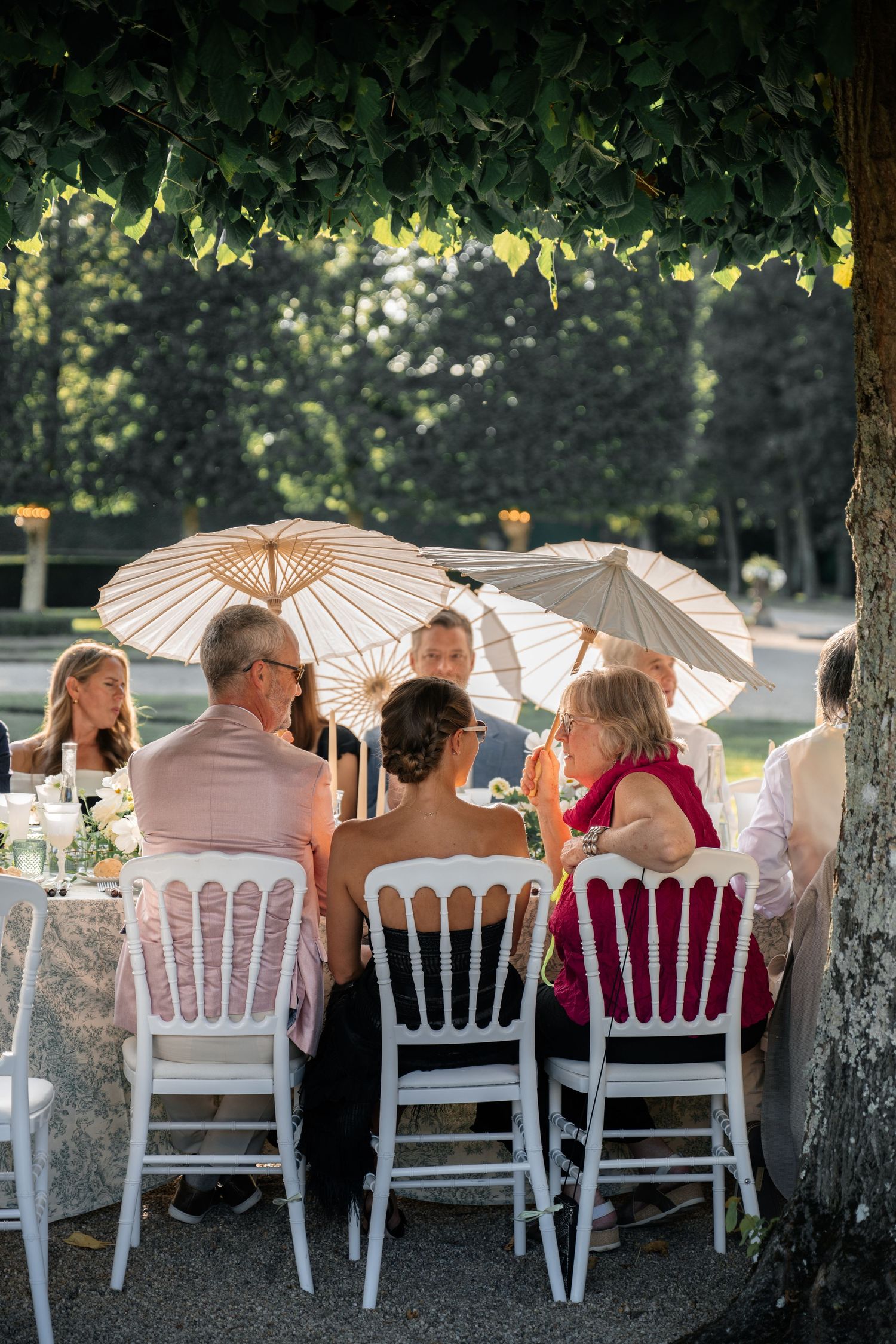 Wedding guests seated at outdoor reception tables under tree canopy with white parasol umbrellas.