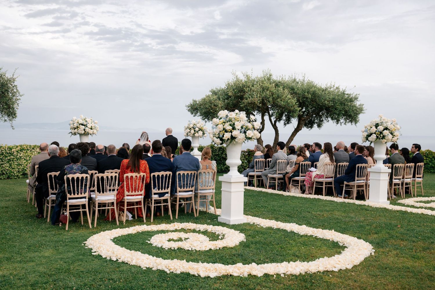 Outdoor wedding ceremony with guests seated in chairs around floral petal spiral aisle by the sea.
