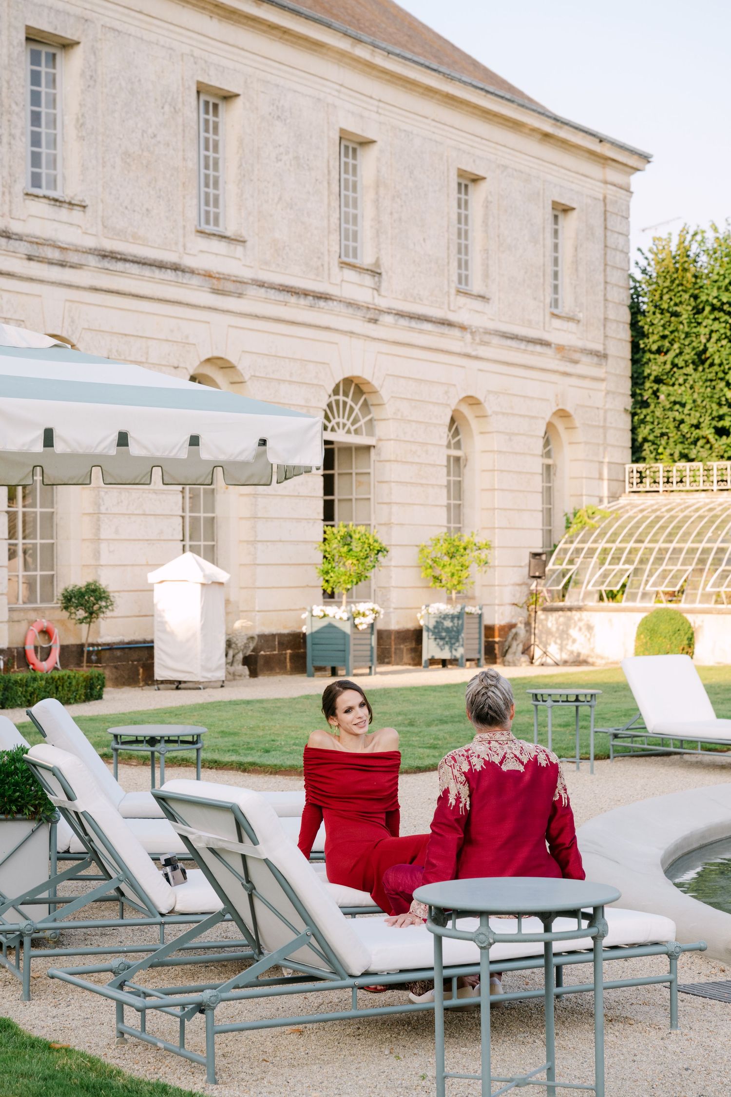 Two women in red dresses conversing on loungers beside pool at grand French chateau venue.