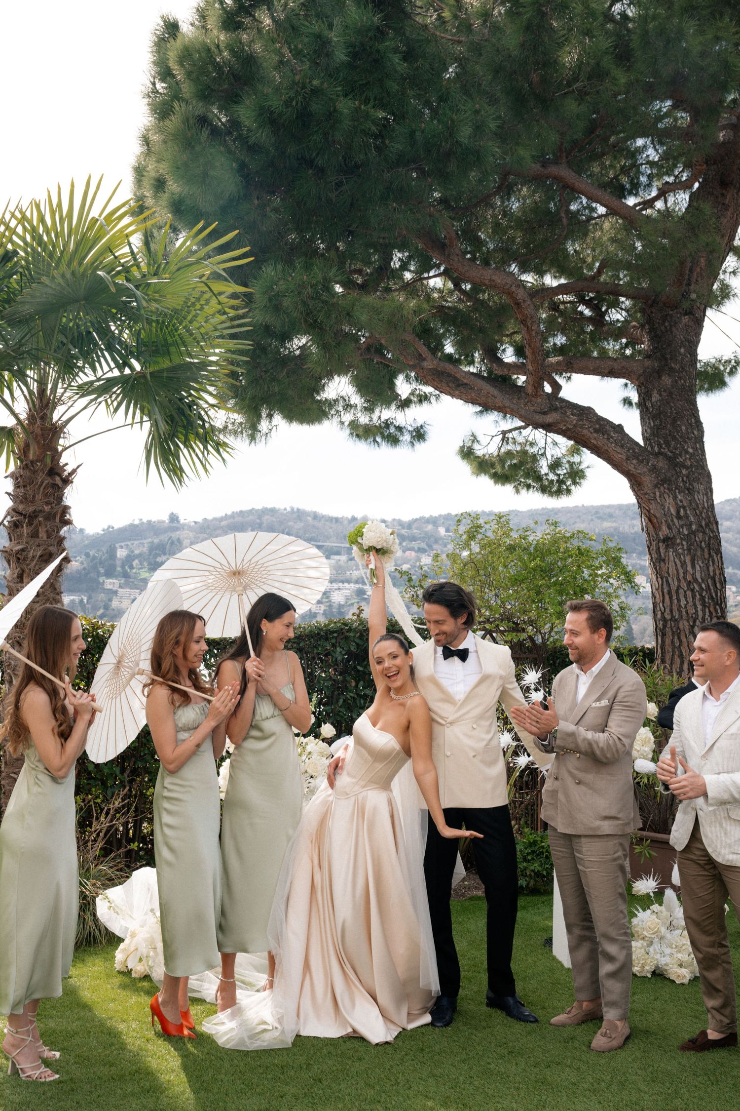Joyful newlyweds celebrate with bridesmaids in sage green dresses holding white parasols at outdoor wedding.