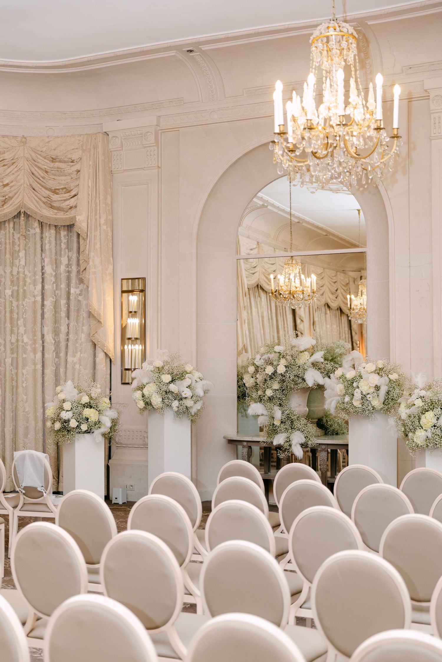 Elegant wedding ceremony venue with white chairs, floral arrangements, crystal chandelier, and arched mirror in a cream ballroom.