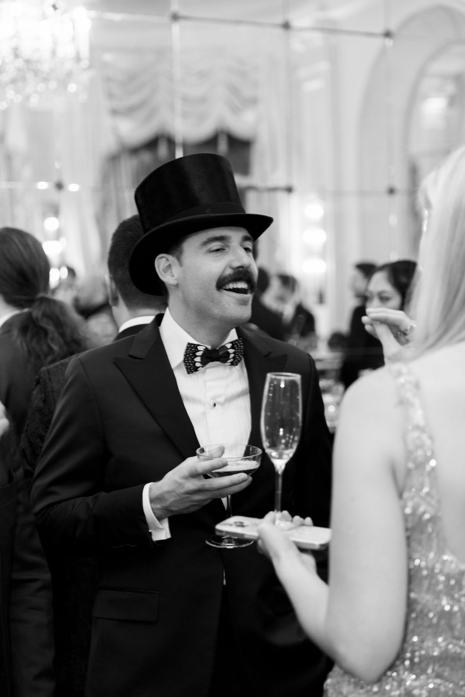 A dapper man in a top hat and bow tie laughs while holding champagne and a small plate at a formal gala.