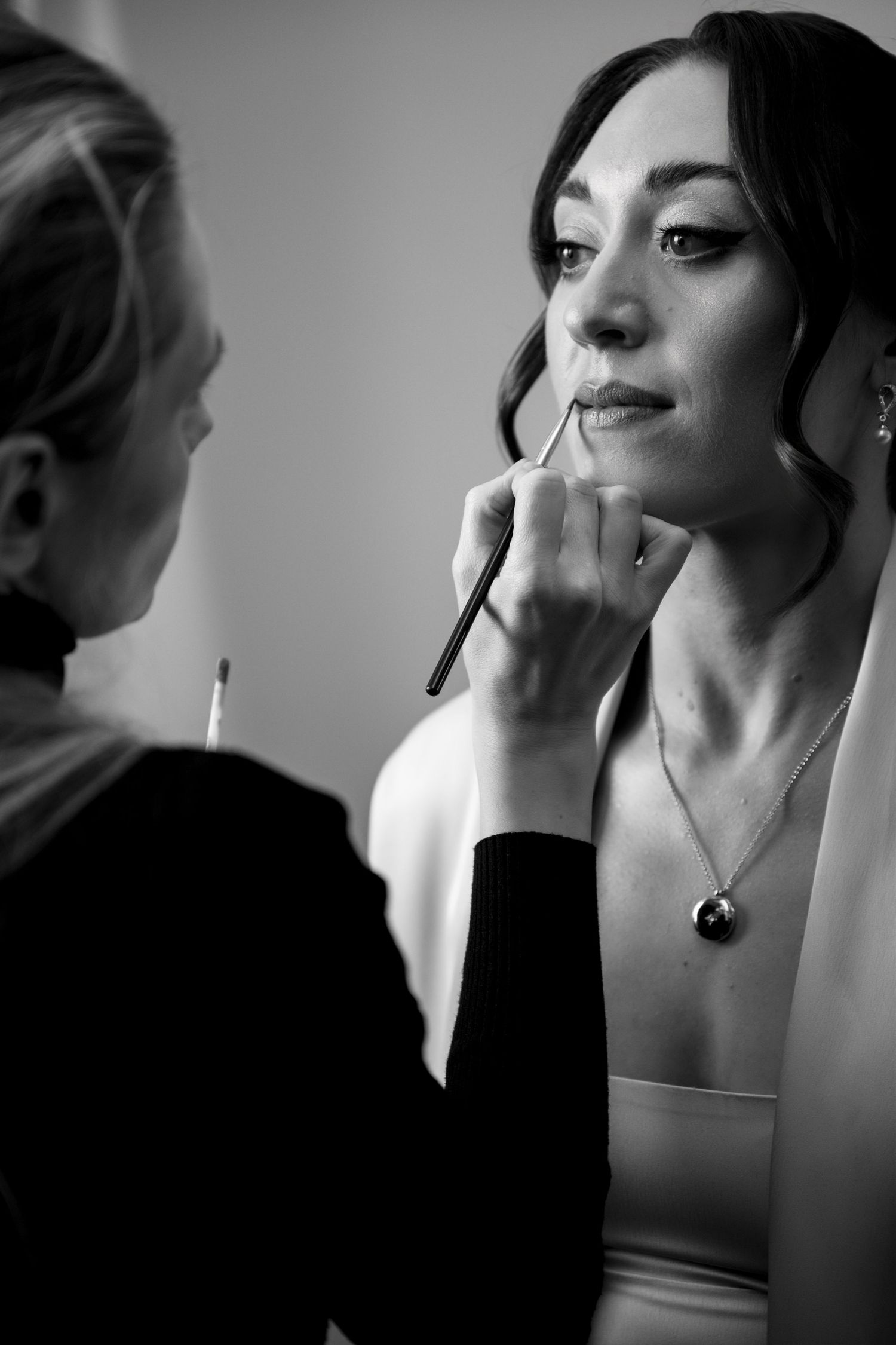 Black and white photo of a makeup artist applying lipstick to a bride wearing a white dress and pearl necklace.