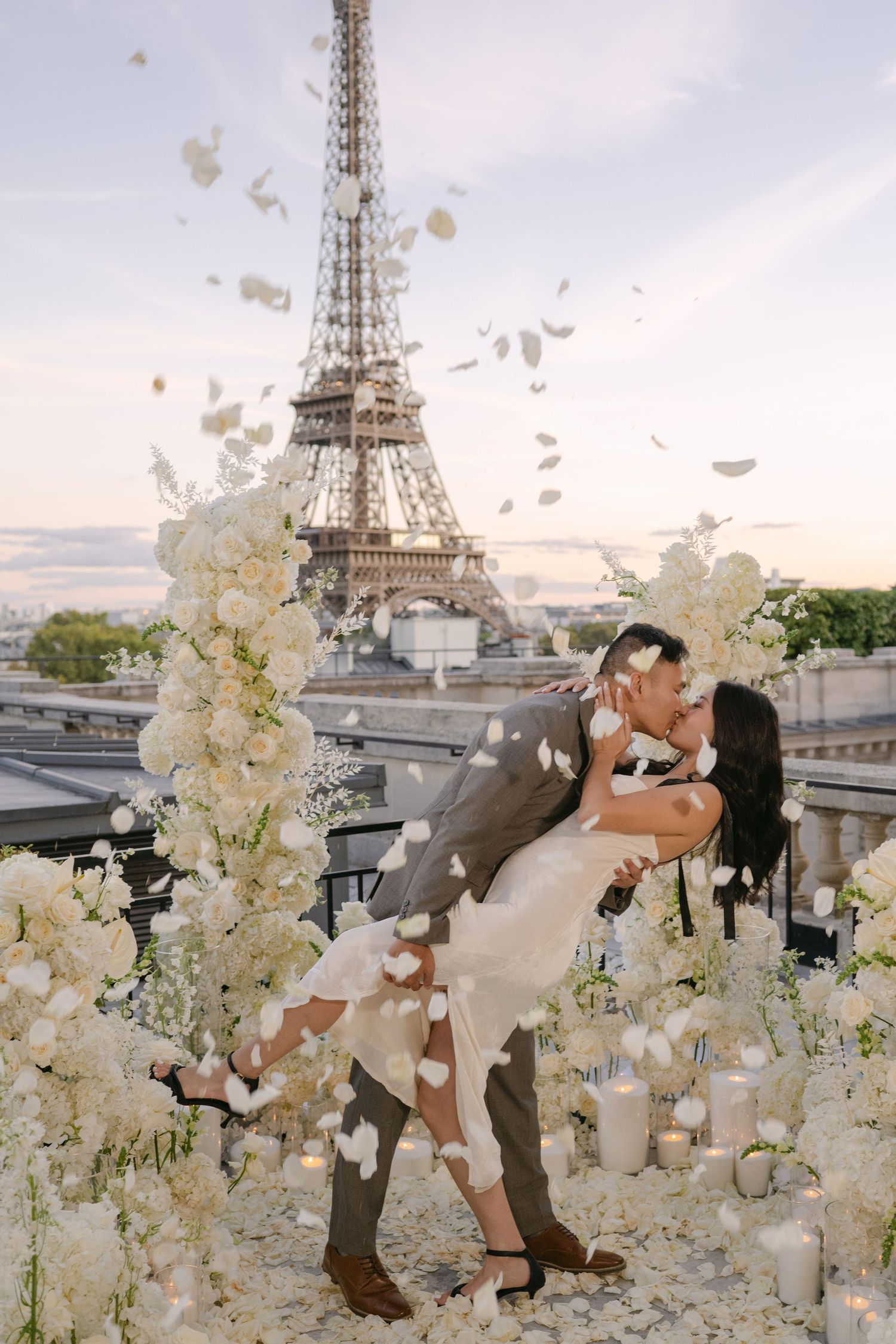 Couple embracing in a romantic dip surrounded by white flower arrangements and petals on a Paris rooftop with the Eiffel Tower at sunset.
