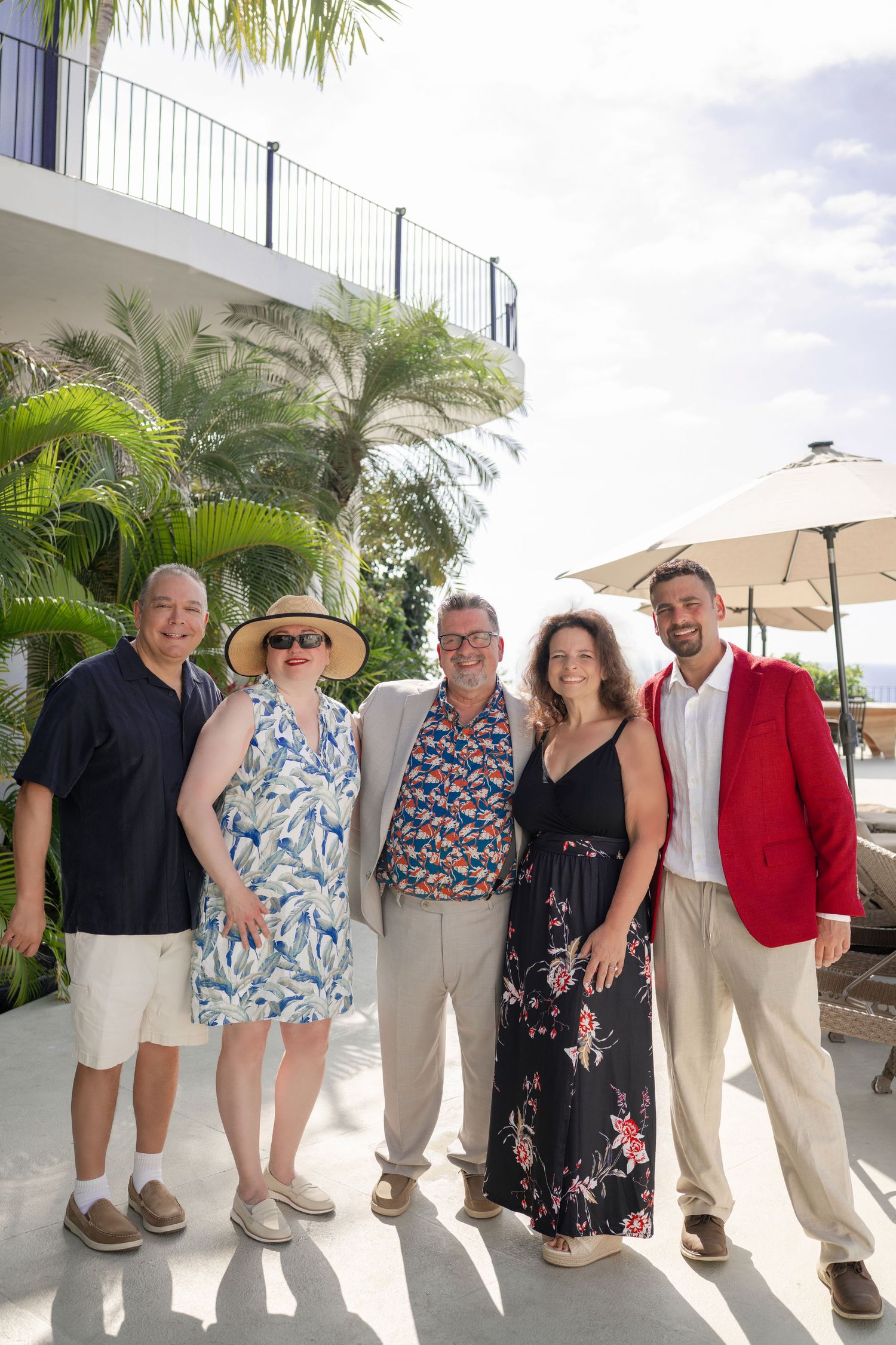 Group of five smiling adults posing together outdoors at a tropical resort with palm trees and umbrella.