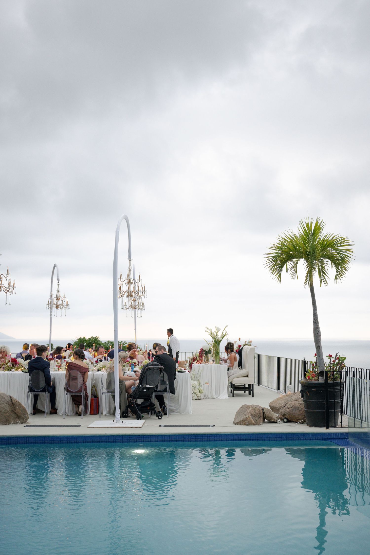Elegant outdoor wedding reception beside a pool with chandeliers, long white tables, and ocean views under cloudy skies.