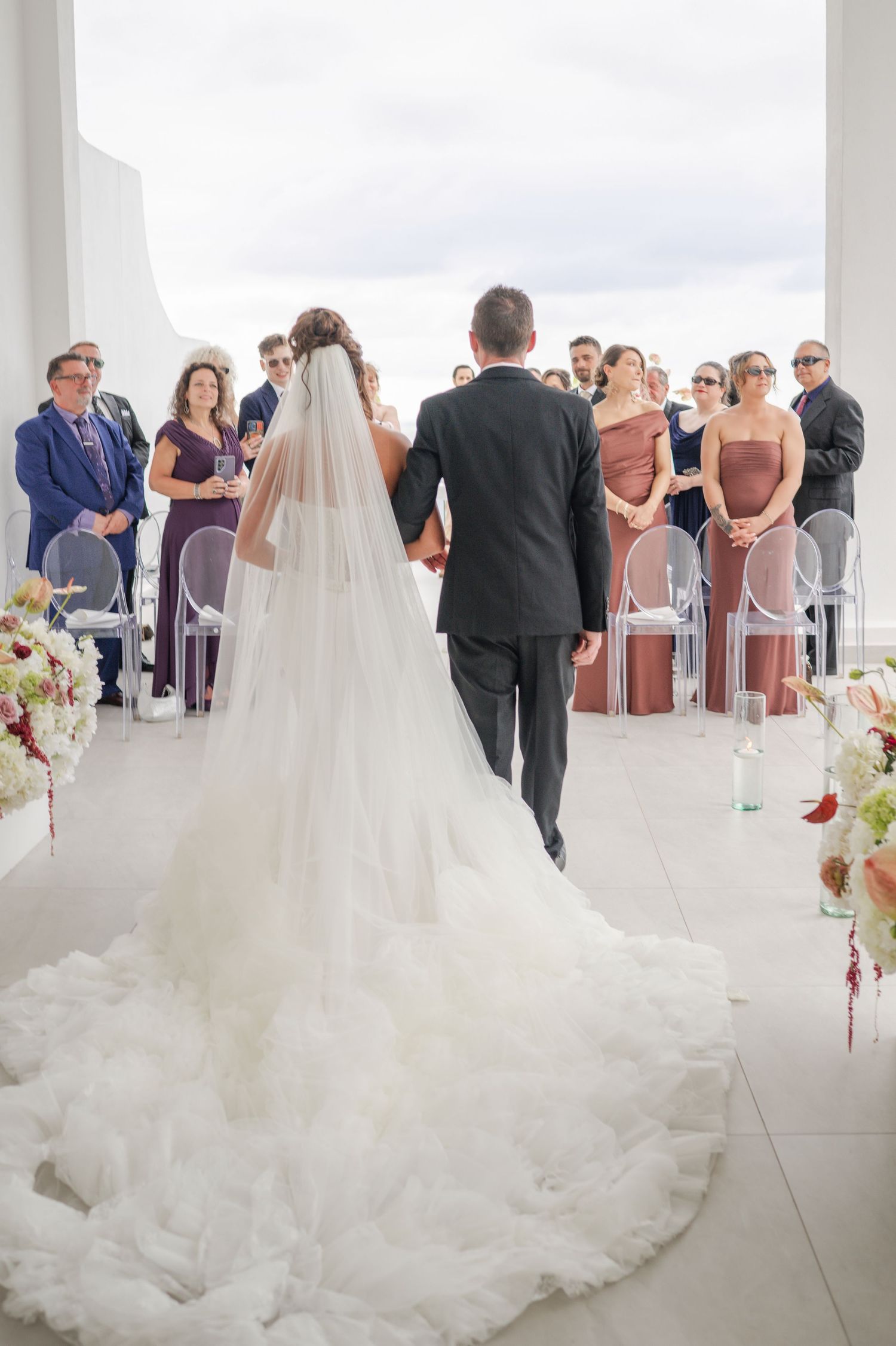 Bride in white gown with veil walks down aisle with groom toward guests at outdoor wedding ceremony.