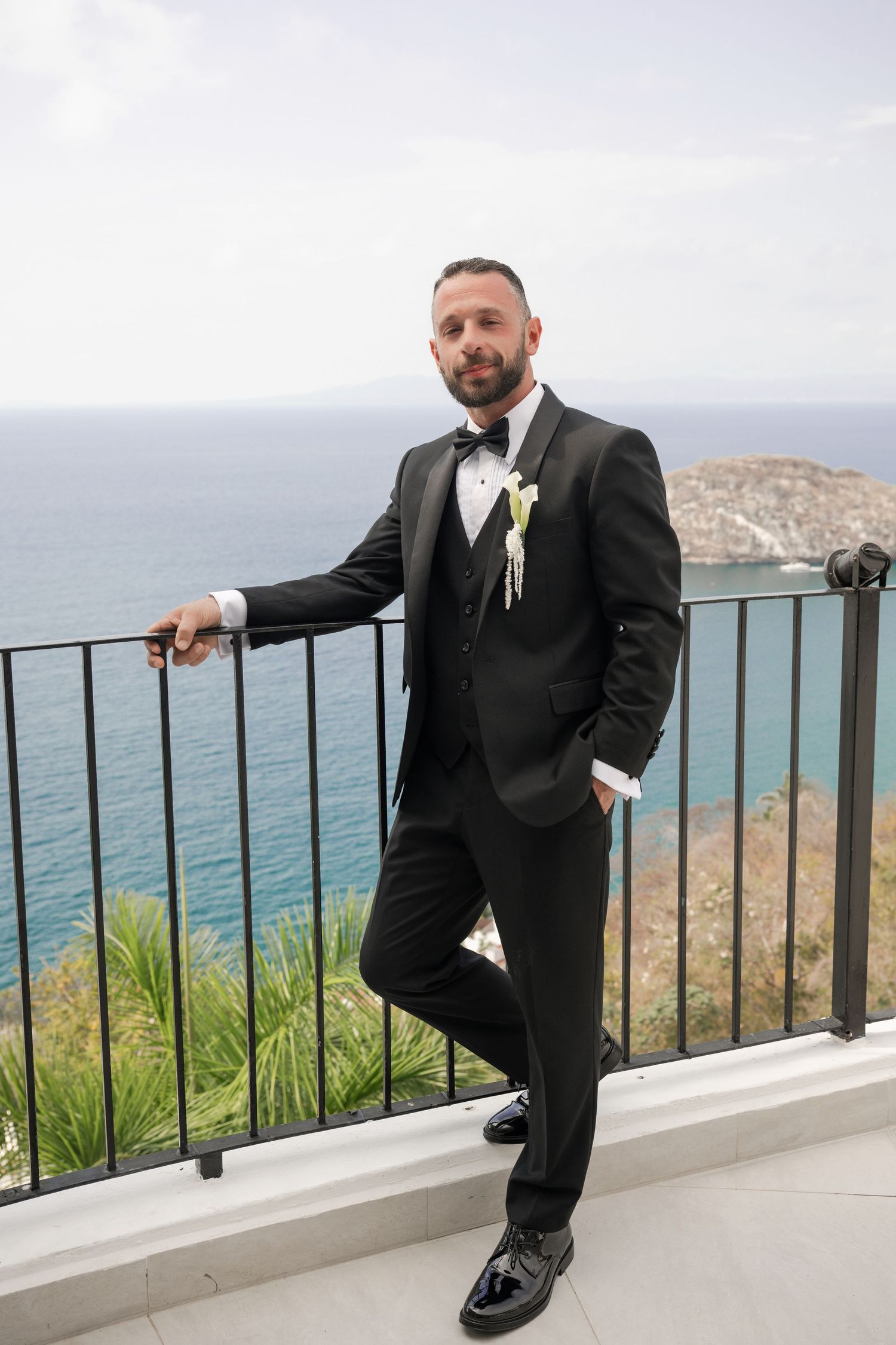 Groom in black tuxedo with boutonniere poses on seaside balcony with ocean view.