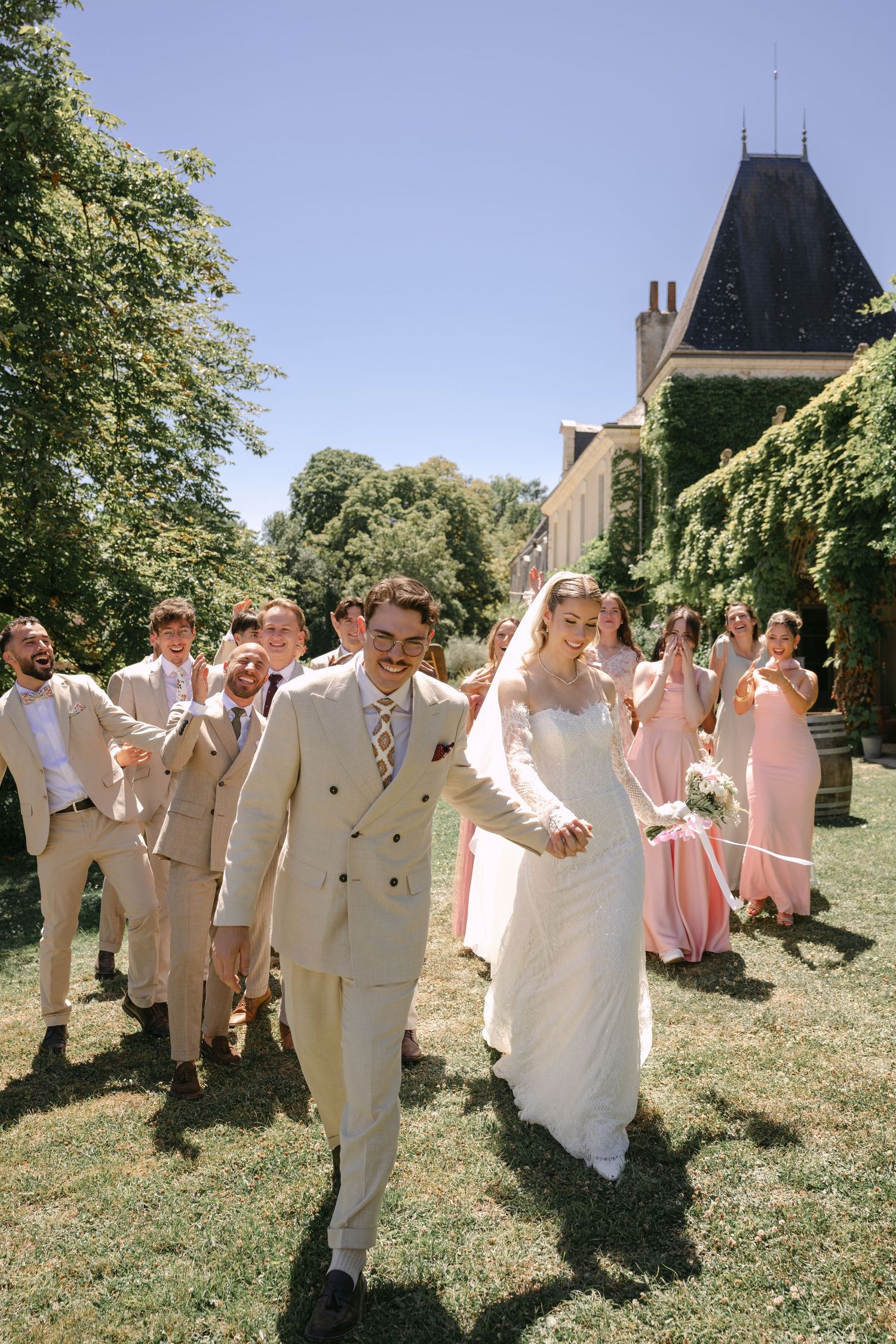 Bride and groom walk joyfully outdoors at a sunny wedding venue, surrounded by cheering bridesmaids in pink and groomsmen.