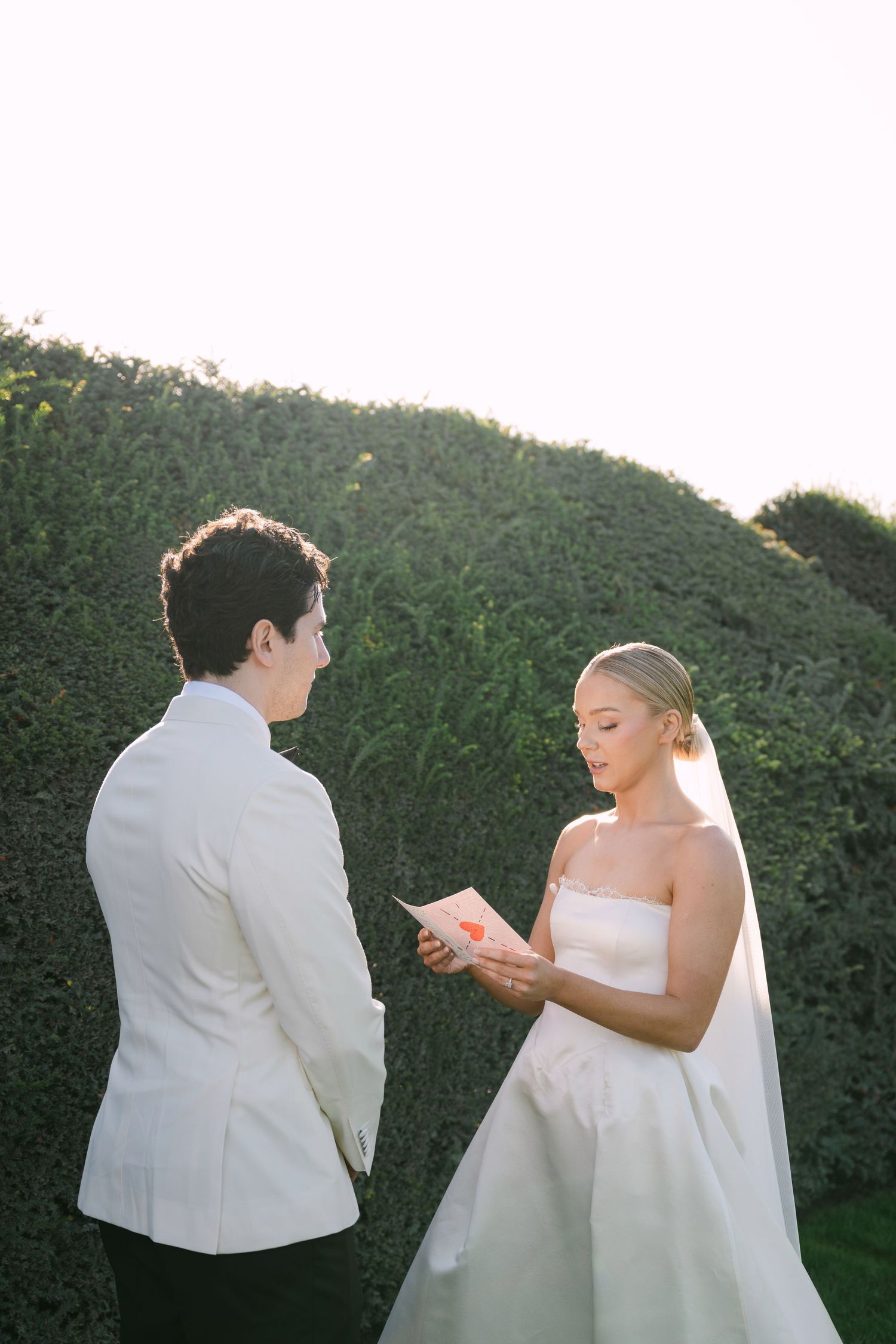 Bride reads wedding vows to groom in white tuxedo jacket outdoors at golden hour ceremony.
