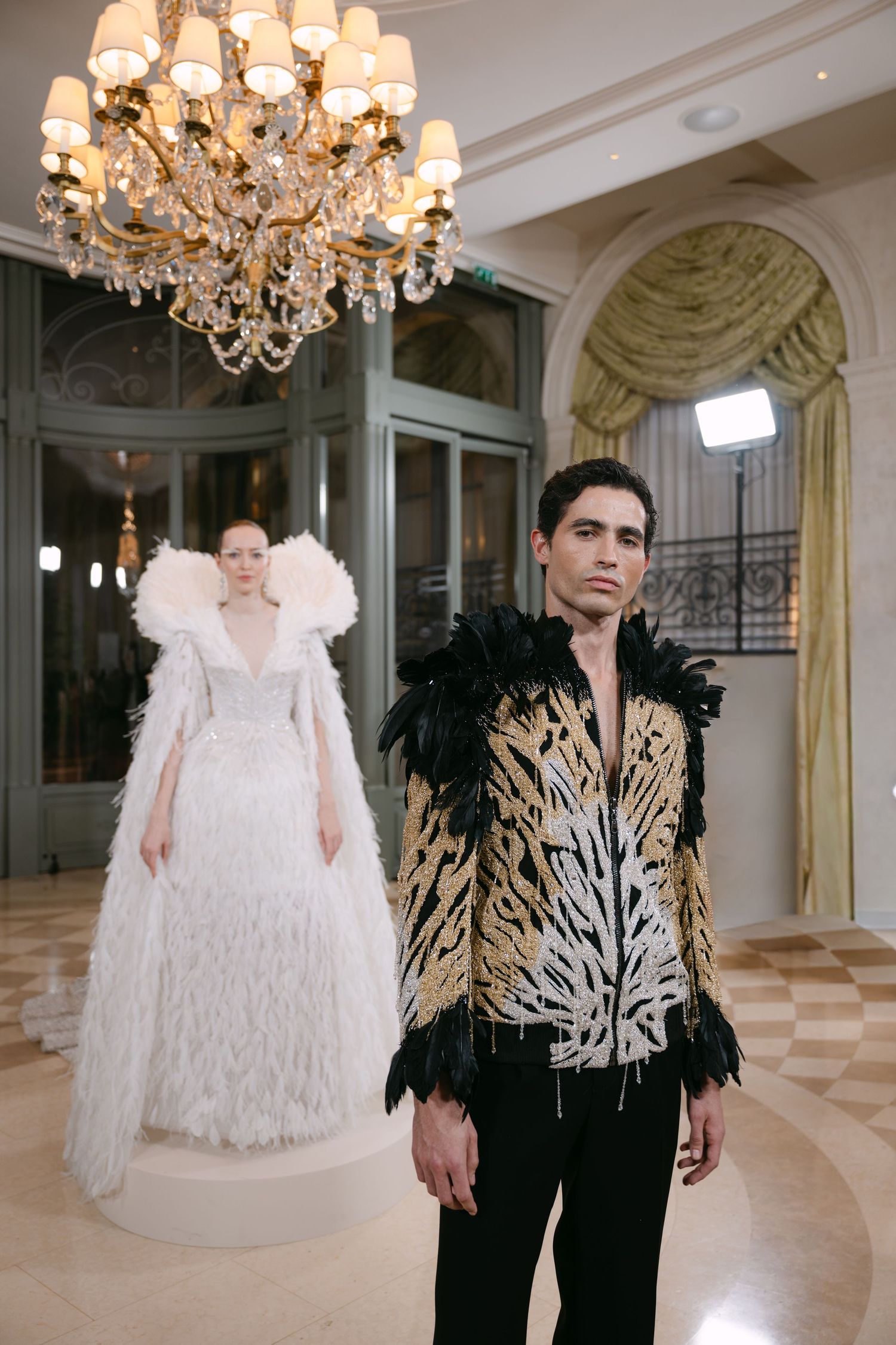 Male model in gold embroidered jacket with black feathers stands before white feathered gown.
