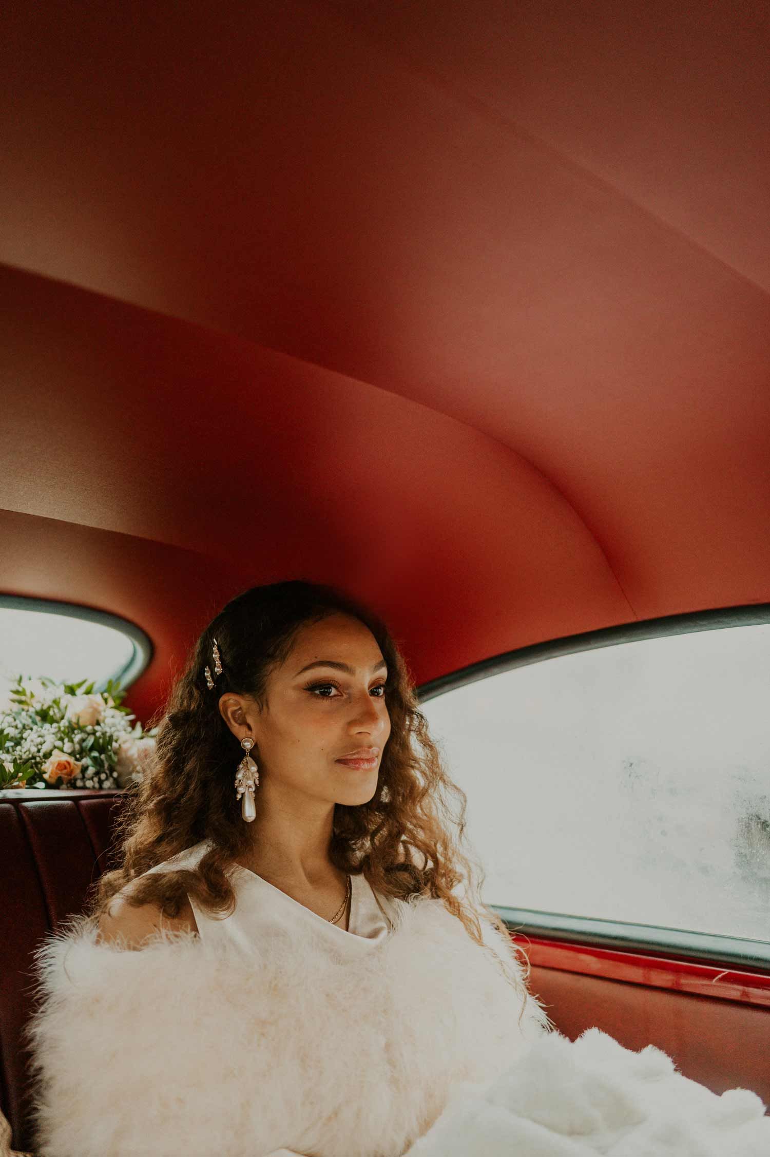 A bride in a white fluffy dress sits elegantly in a classic car with red interior.