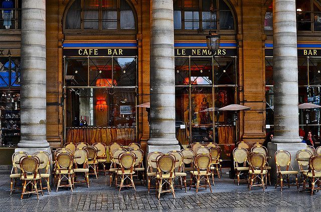 Classic Parisian cafe with rattan chairs and arched stone architecture along a covered walkway.