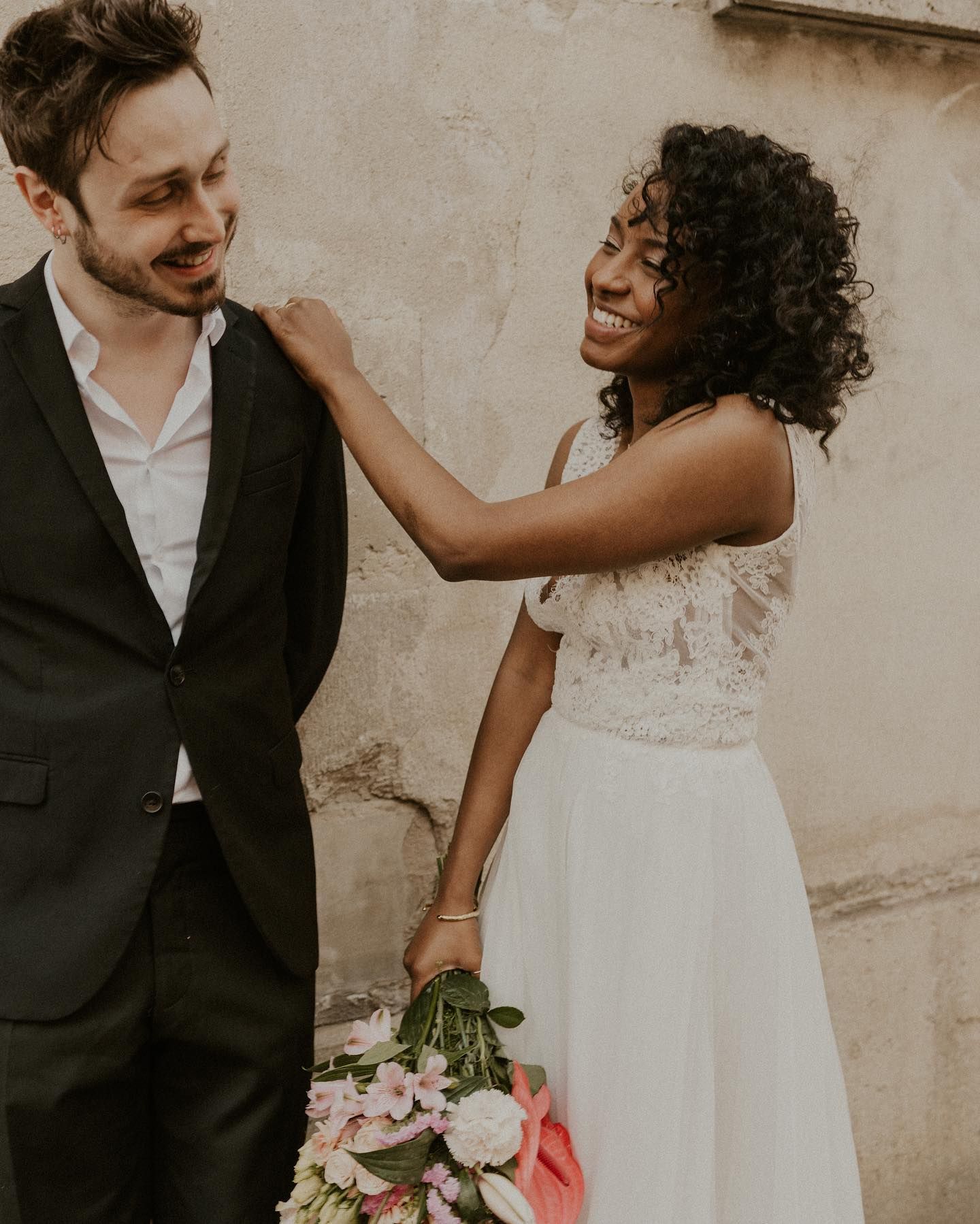 Joyful wedding couple moment against a neutral wall with pink floral bouquet.