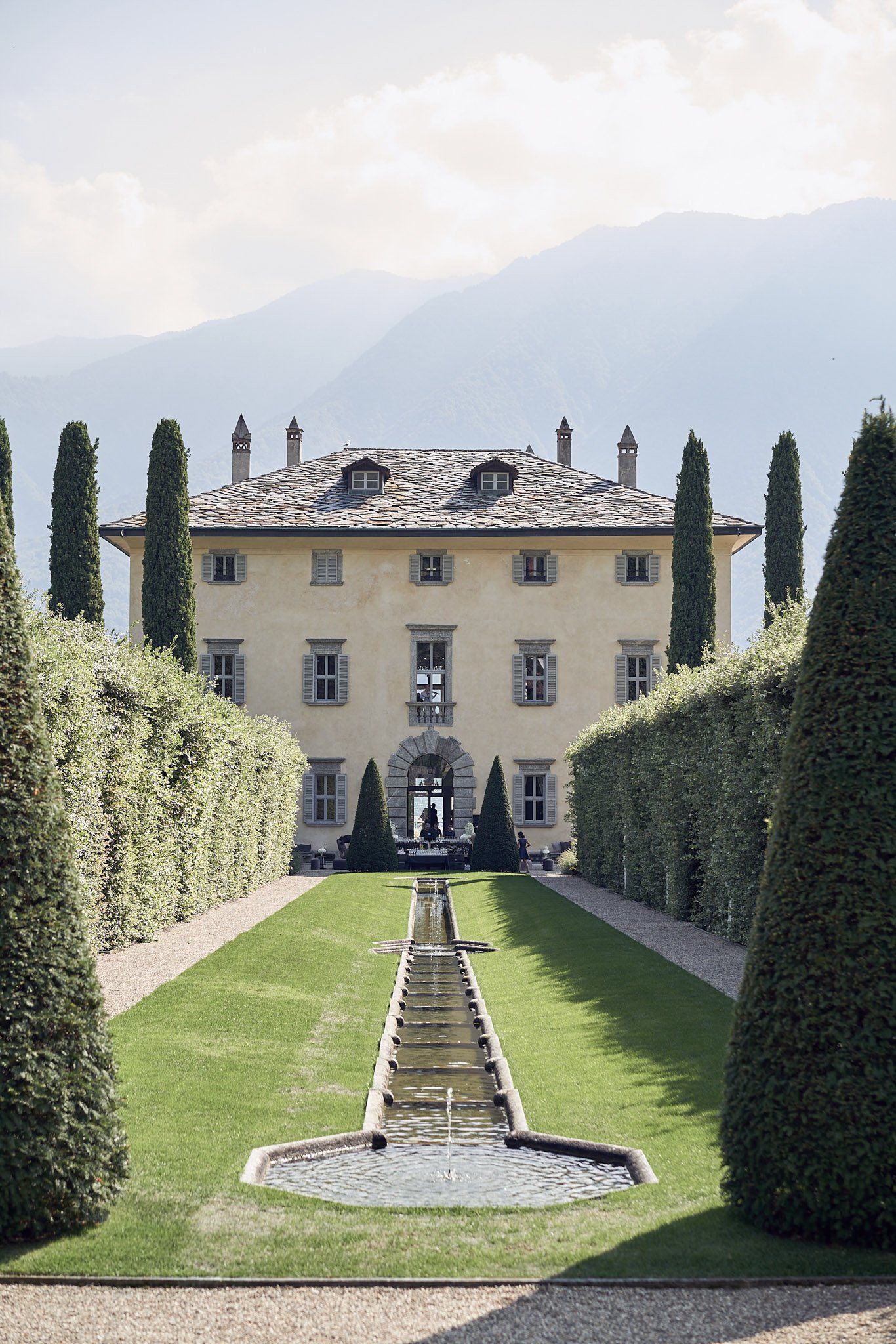 A grand Italian villa with cypress trees and formal garden featuring a water channel leading to the entrance.