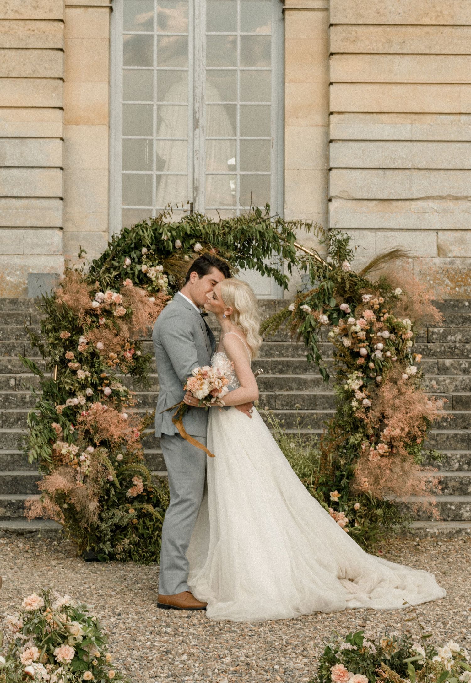 A wedding couple embraces under a romantic floral arch on stone steps of a historic building.