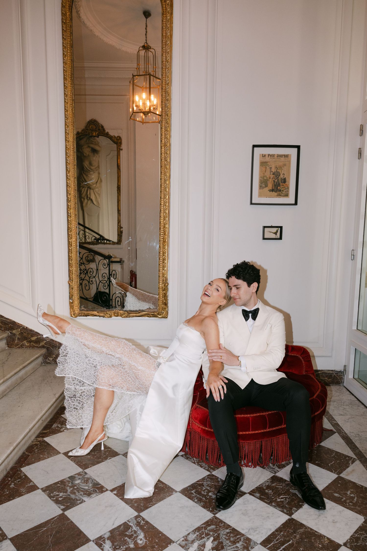 A couple in formal attire poses playfully on a checkered marble floor in an elegant room with ornate gold mirrors.