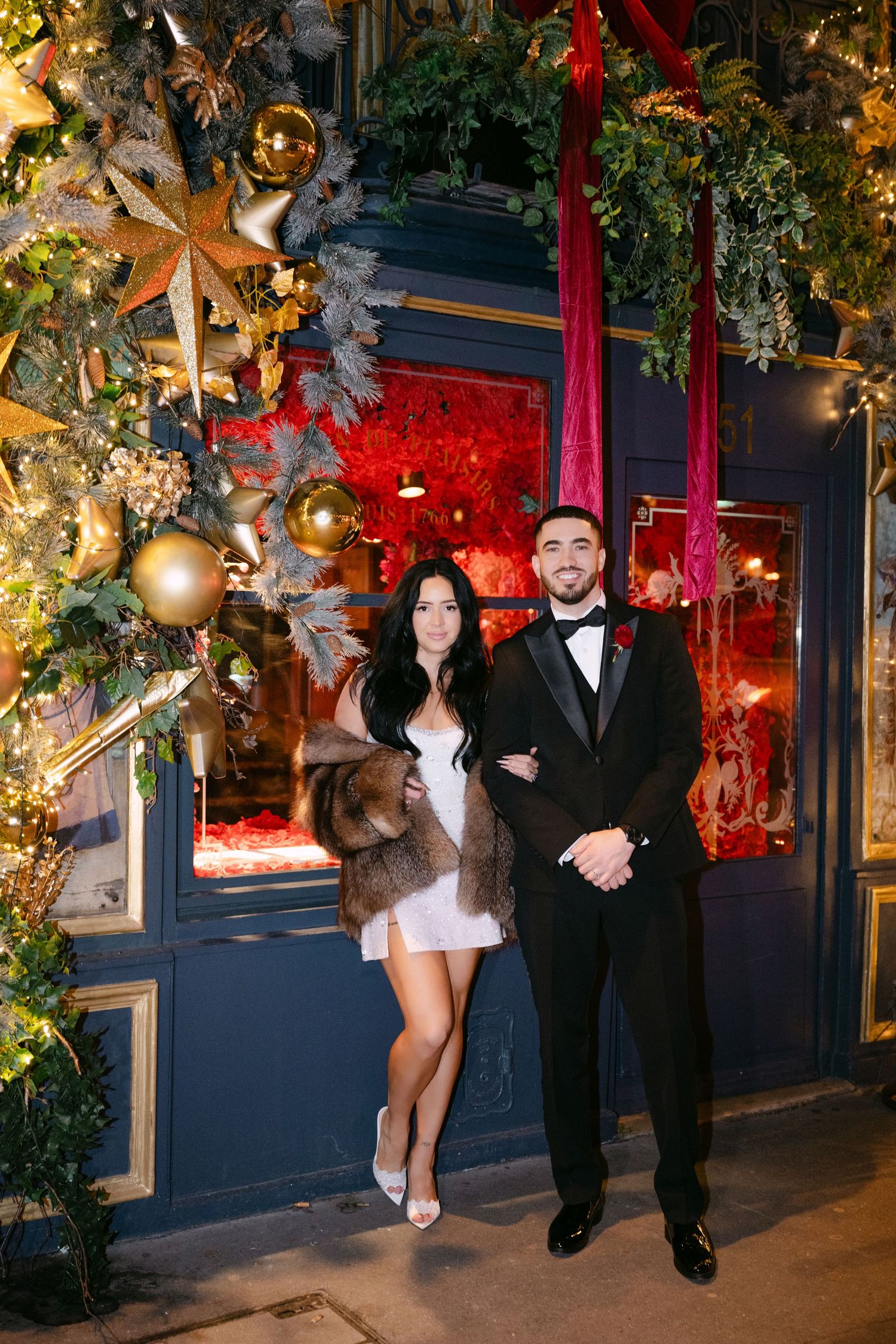 Elegant couple in formal attire poses before festive Christmas decorations outside Parisian restaurant.