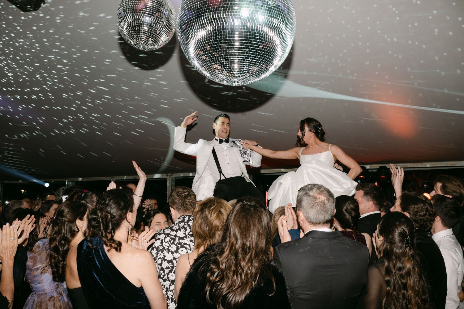 Bride and groom lifted by guests on crowded wedding dance floor under disco ball.