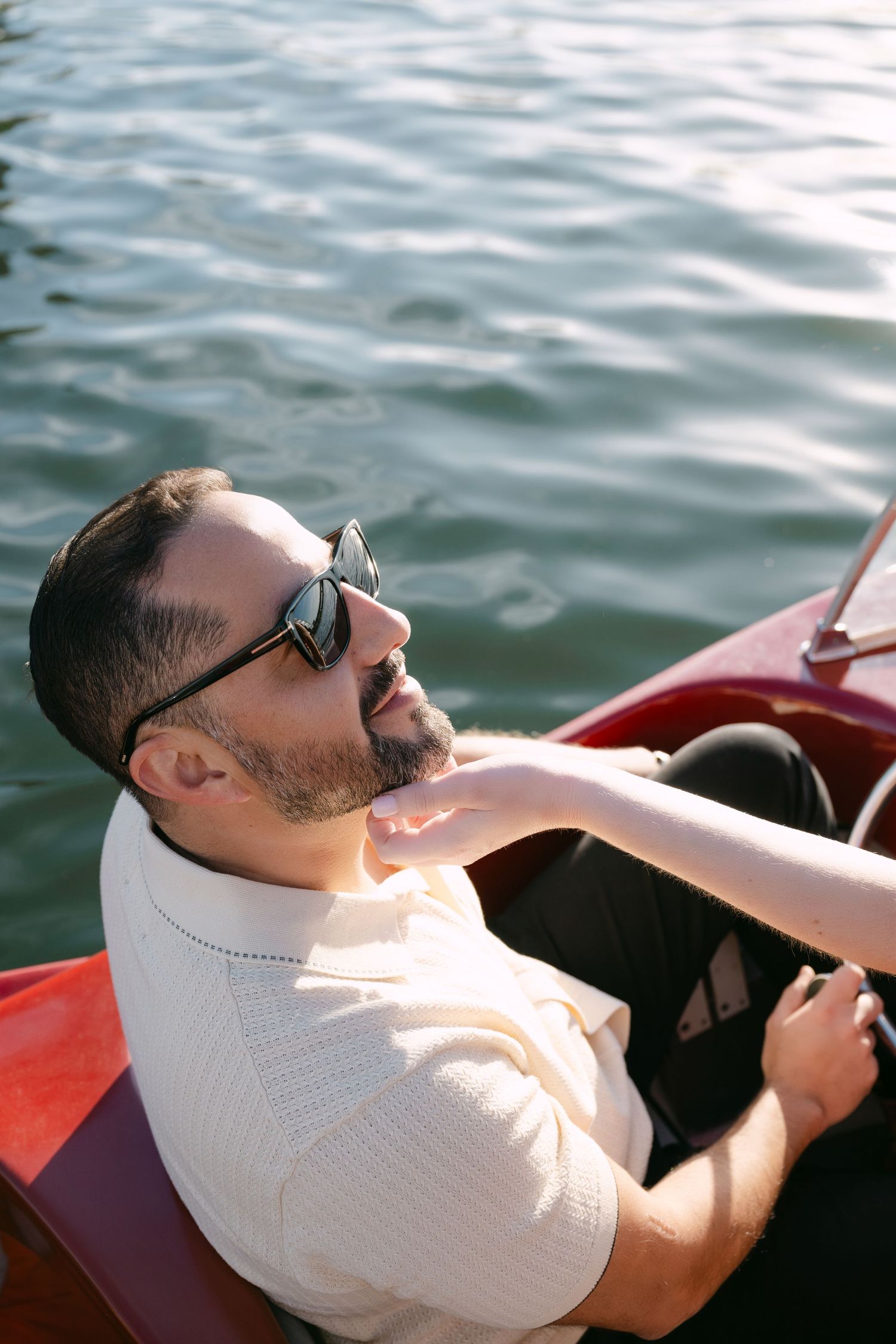 Man with beard and sunglasses relaxing on a red boat, hand gently touching his neck.