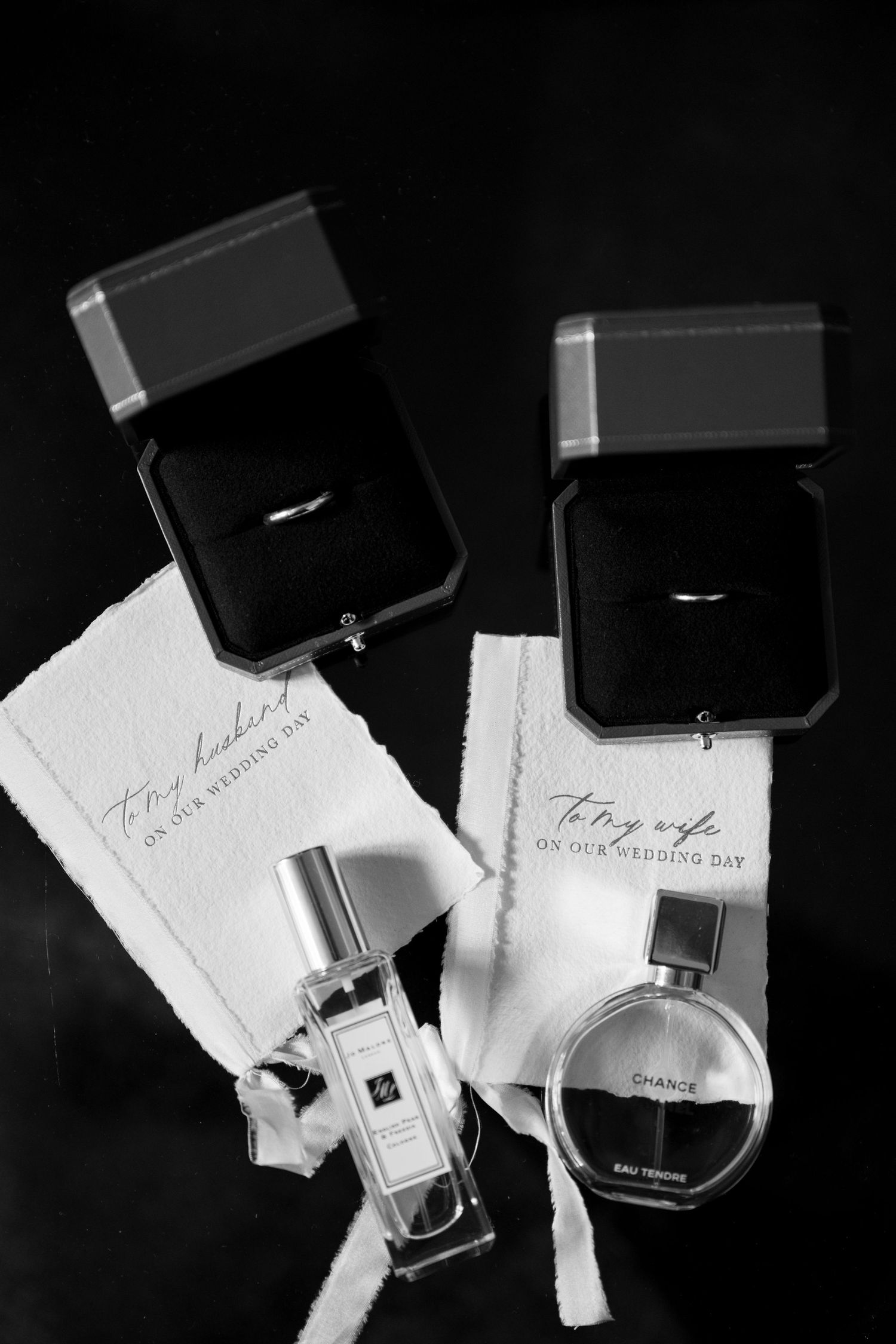 Black and white flat lay of wedding rings in boxes with perfume bottles and wedding day cards.