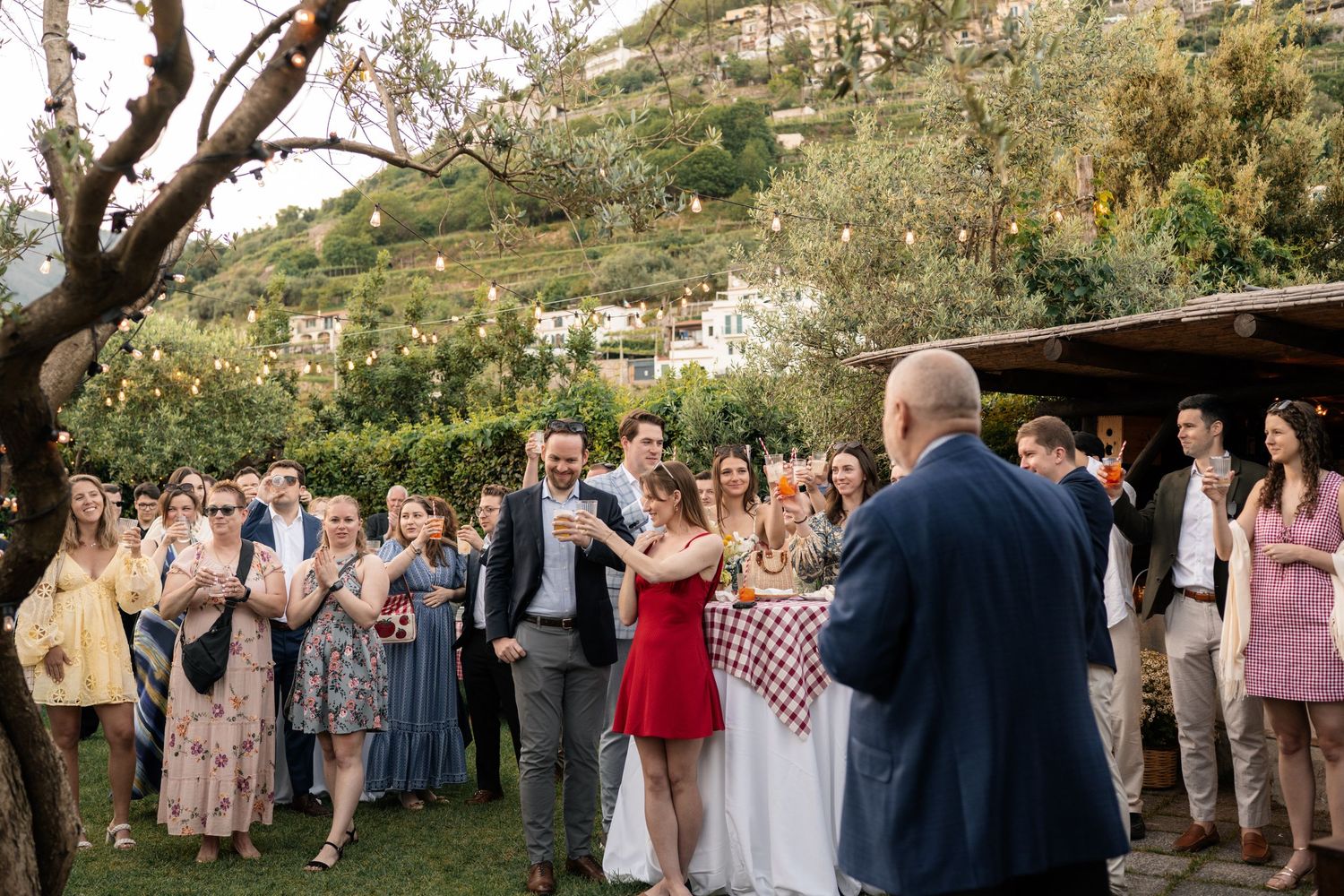 Wedding guests applaud a couple at an outdoor Italian garden reception with string lights and hillside views.