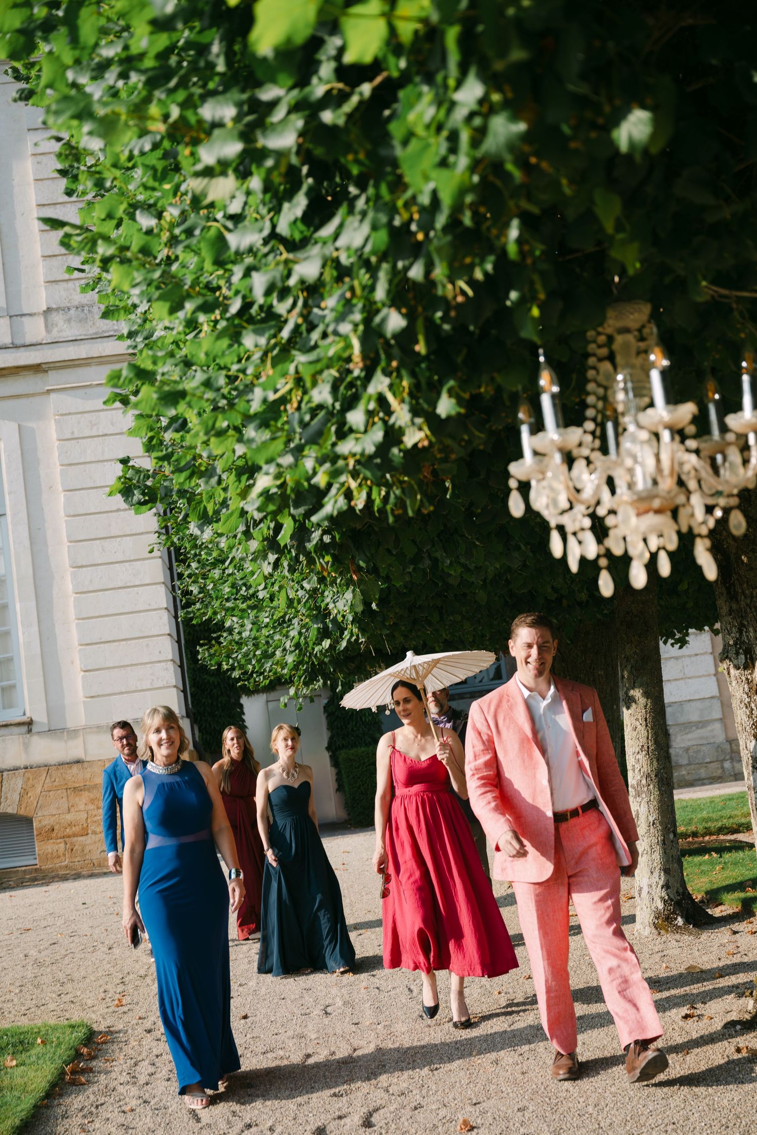 Wedding guests in colorful formal attire walk past a hanging crystal chandelier beneath a canopy of green trees.