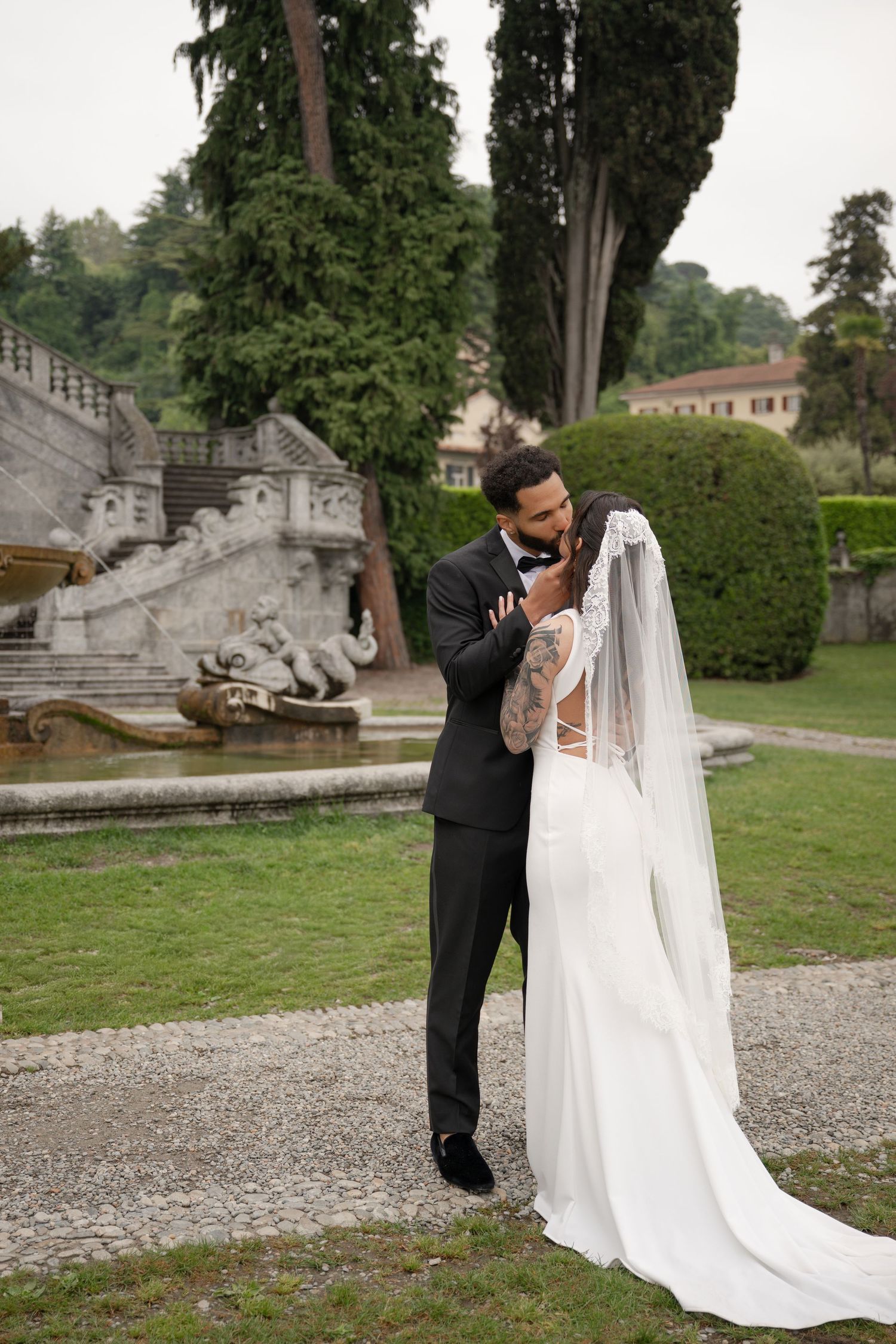 Groom in black tuxedo embracing bride in white gown near ornate fountain in lush garden.