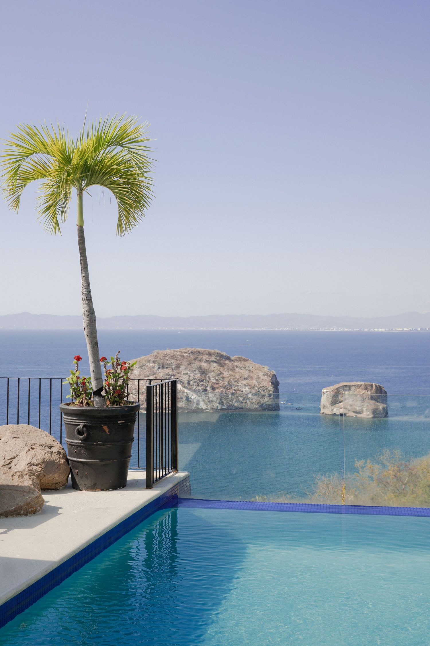 Infinity pool overlooking blue ocean with palm tree, rocky outcrops, and clear sky at a luxury resort.