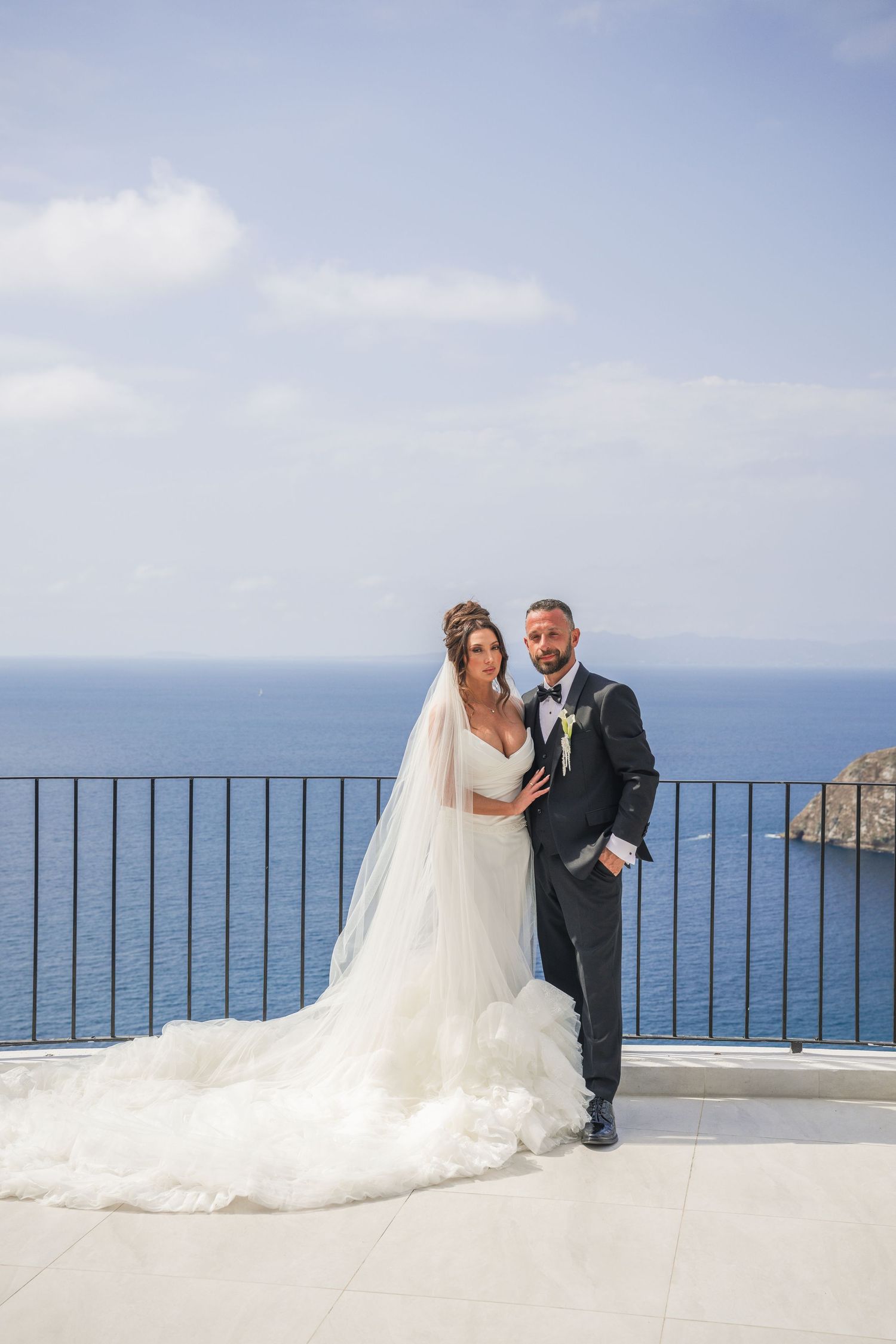Bride in white gown with veil and groom in black suit pose on terrace with ocean backdrop on wedding day.